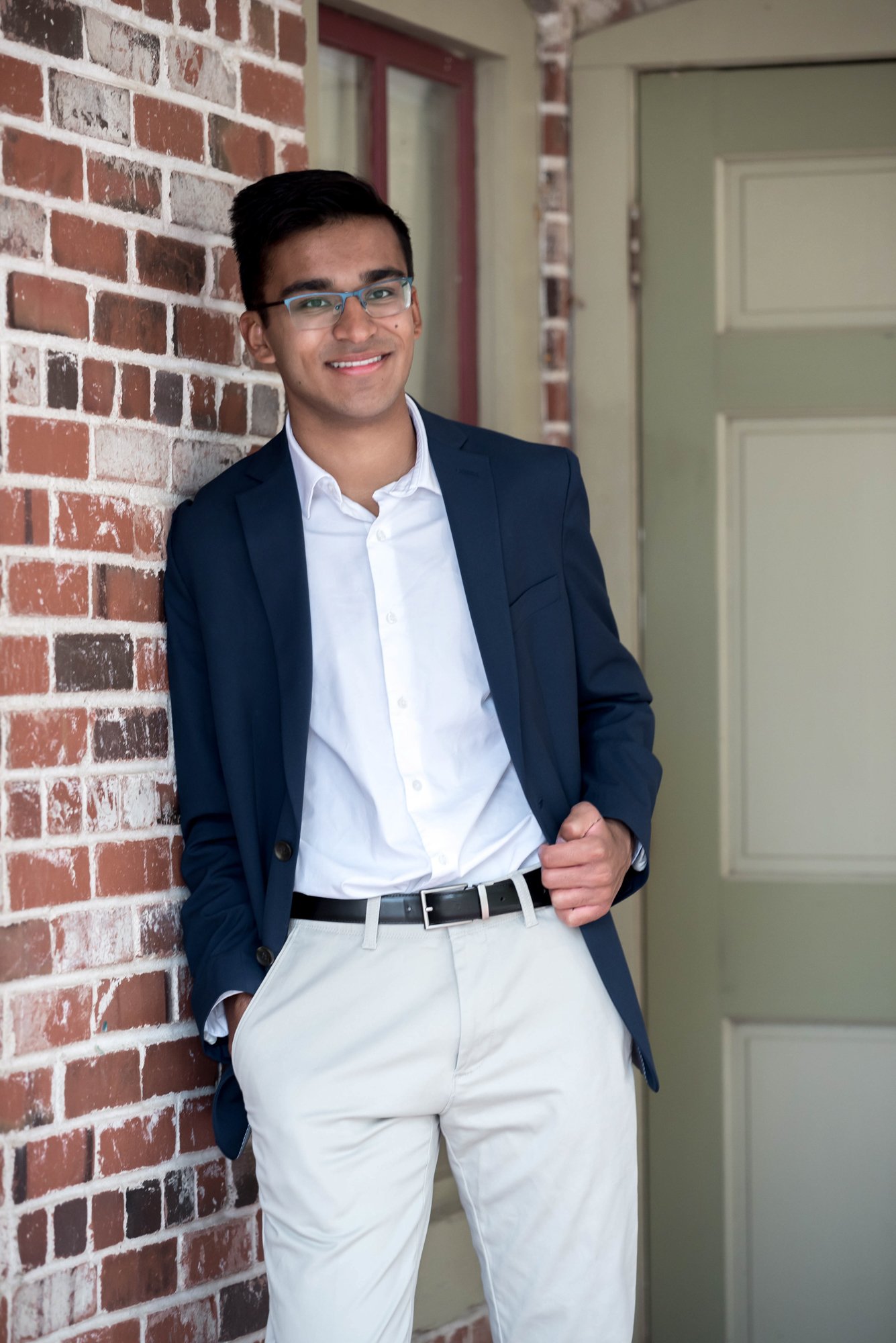 Sophisticated senior photo of a guy wearing a navy blue blazer and glasses, leaning against a rustic brick wall while holding the bottom of his suit jacket at Faust Park in St. Louis.