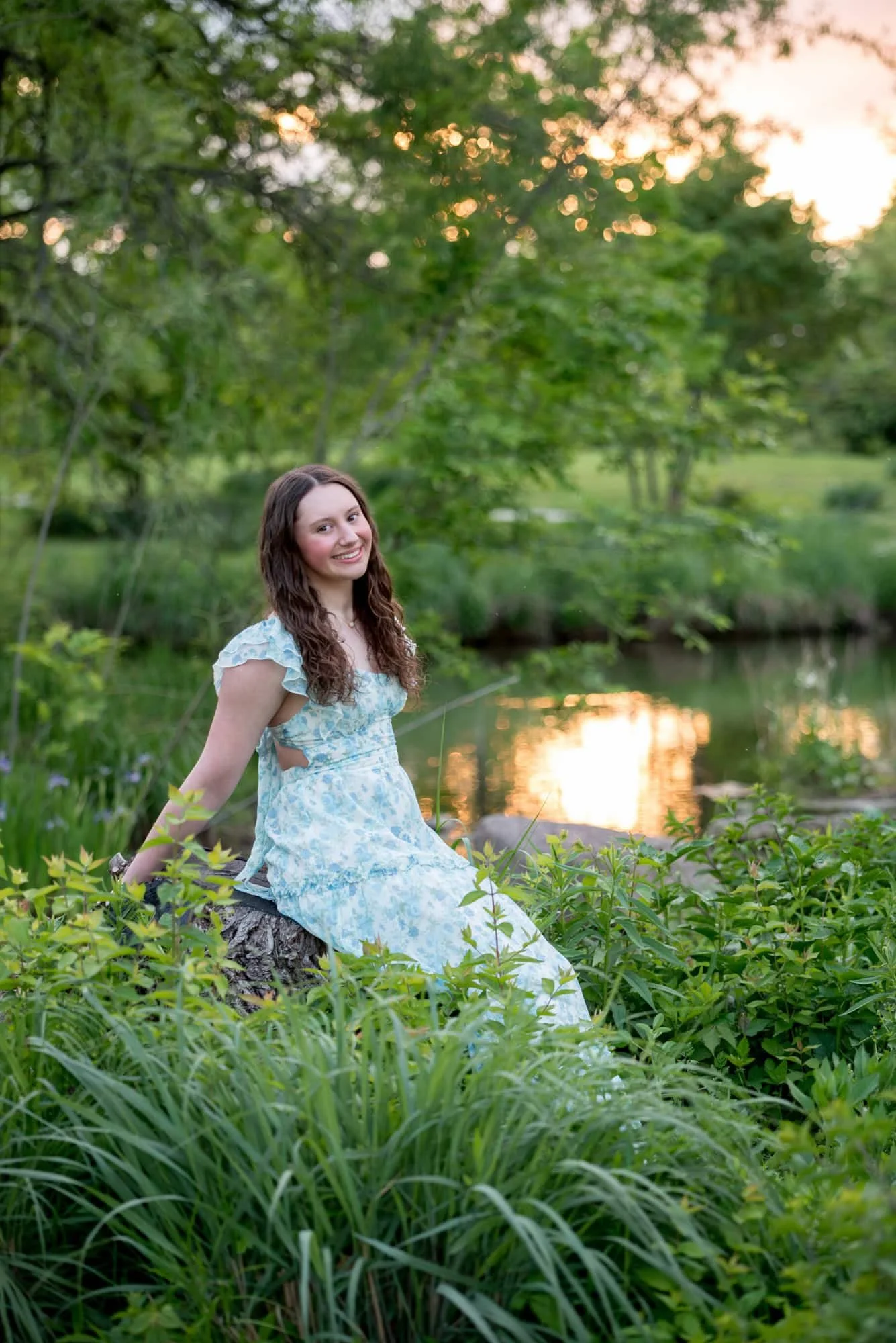 Sunset light reflects on the water behind a high school senior girl sitting in a lush, green creekside meadow in Forest Park.