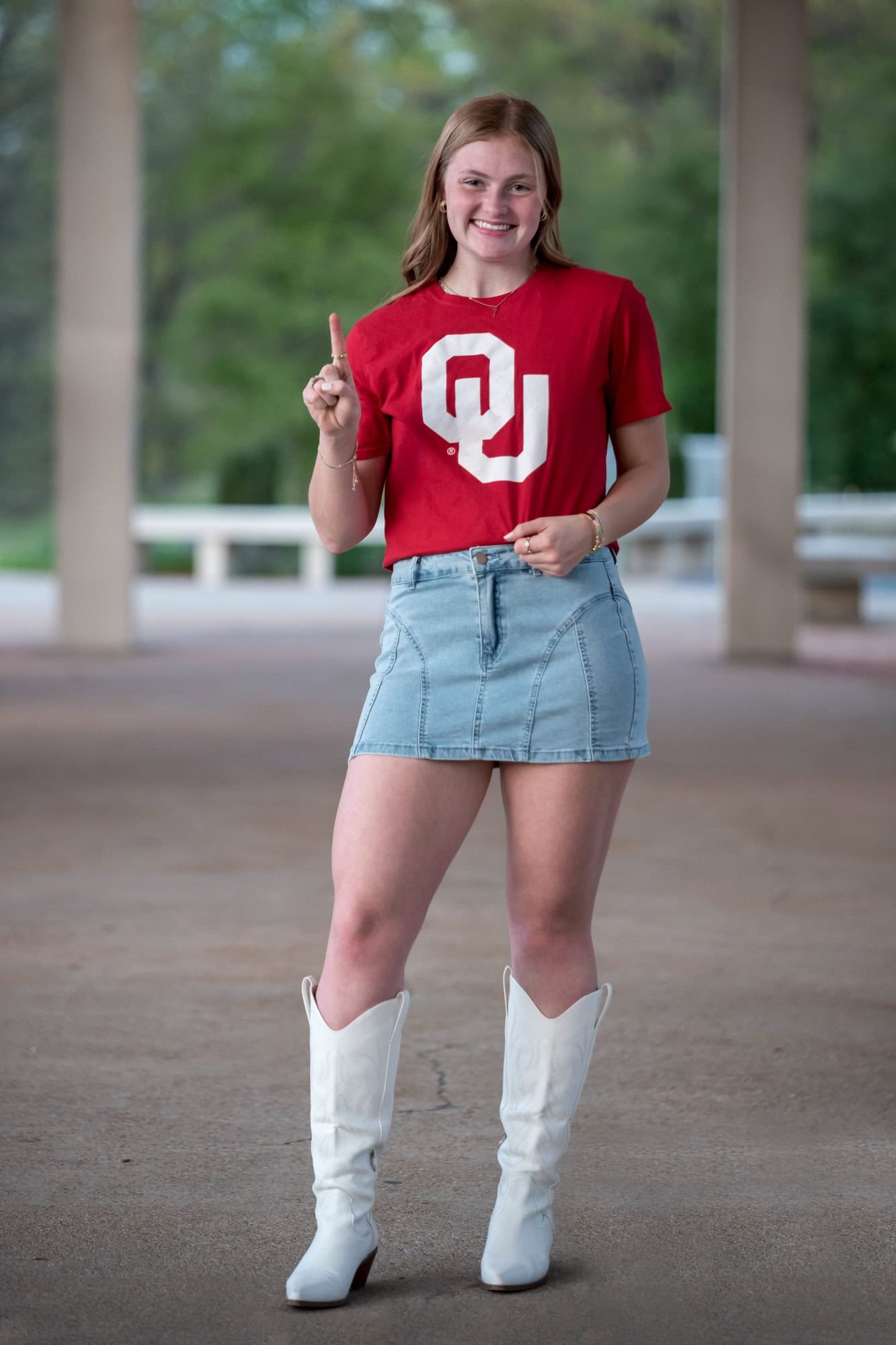 Standing on the open walkway at The Muny in her red OU shirt, denim skirt, and white boots, a senior girl poses for a picture.