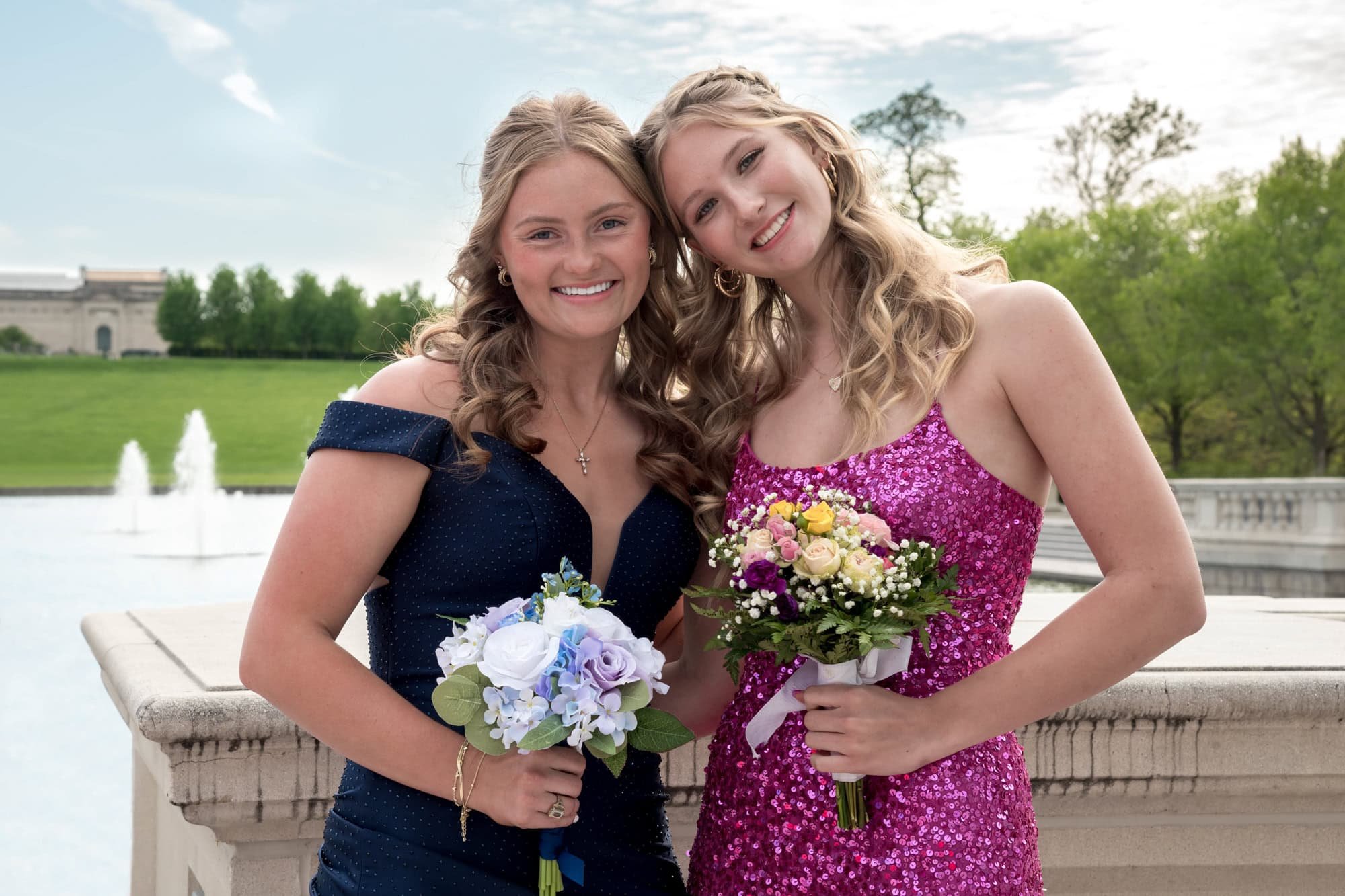 Posing next to a friend in a pink sparkle dress, a senior girl in a navy blue prom dress smiles for a photo in St. Louis County.