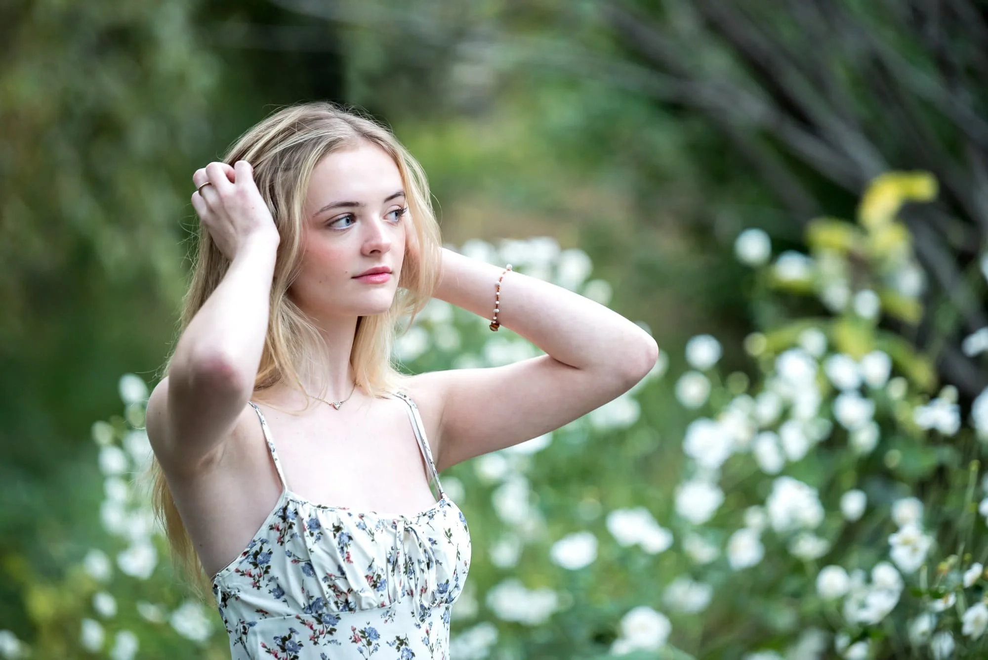 A high school senior girl wearing a white dress with blue flowers stands with her hands in her hair during a fall photo shoot at Lafayette Park.
