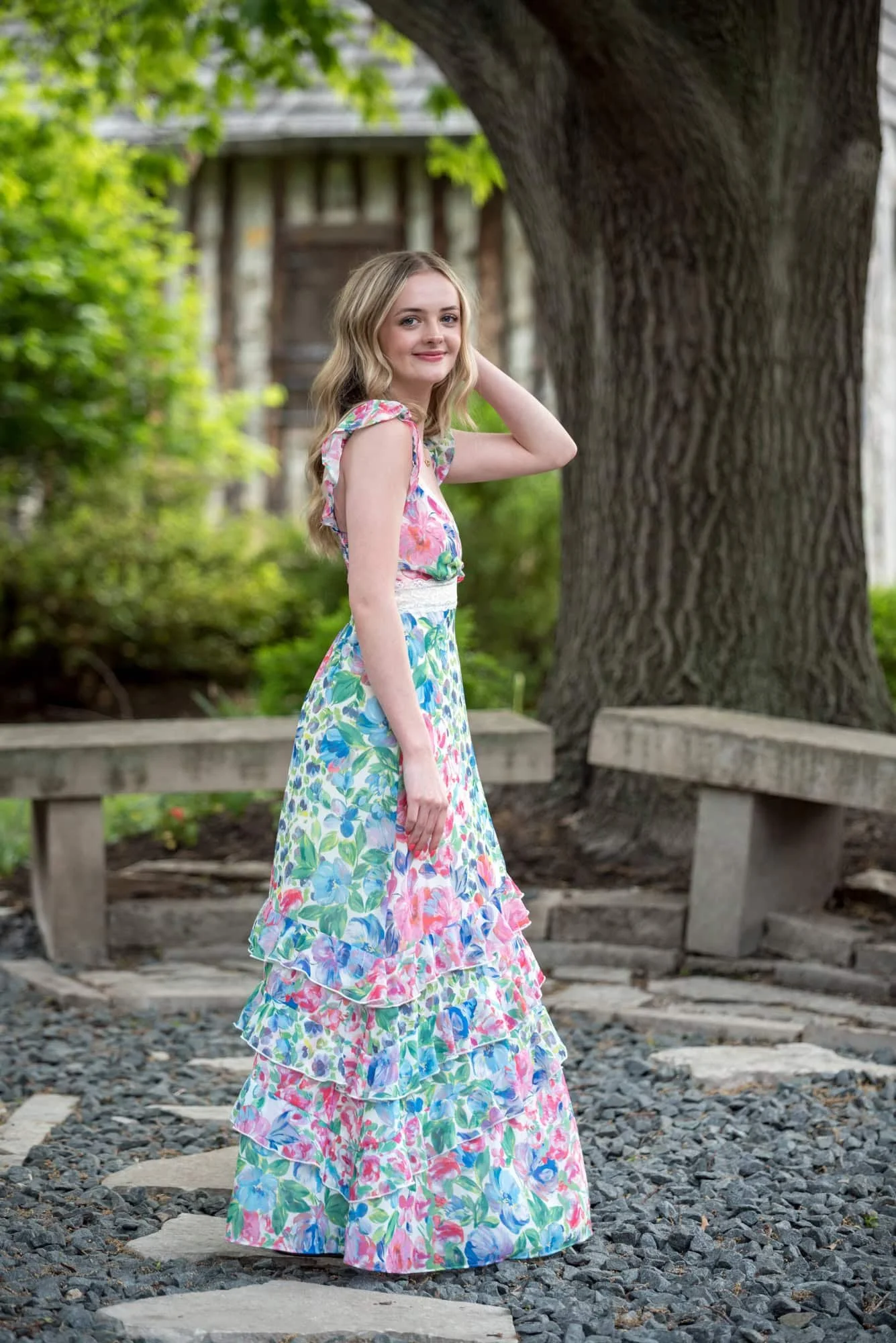 A picture captures a senior girl wearing a floral long dress standing on a stone pathway in front of a historical cabin in St. Charles, MO.