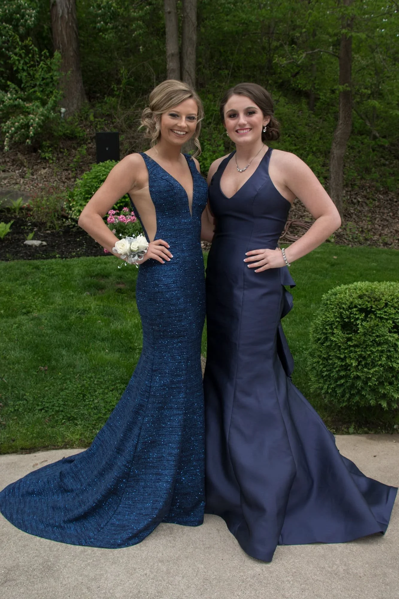 Outdoor photo of two friends smiling together in their elegant prom gowns, one in a sparkling textured navy blue dress and the other in a smooth navy satin gown, taken in Columbia, IL.