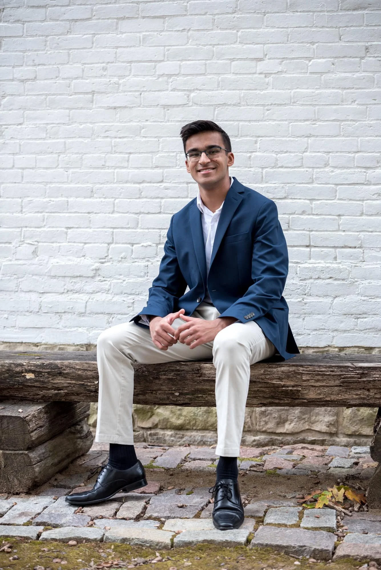 Polished senior photo of a guy in a navy blazer sitting on a wooden bench with his hands clasped together in front of a white brick wall at Faust Park in St. Louis.