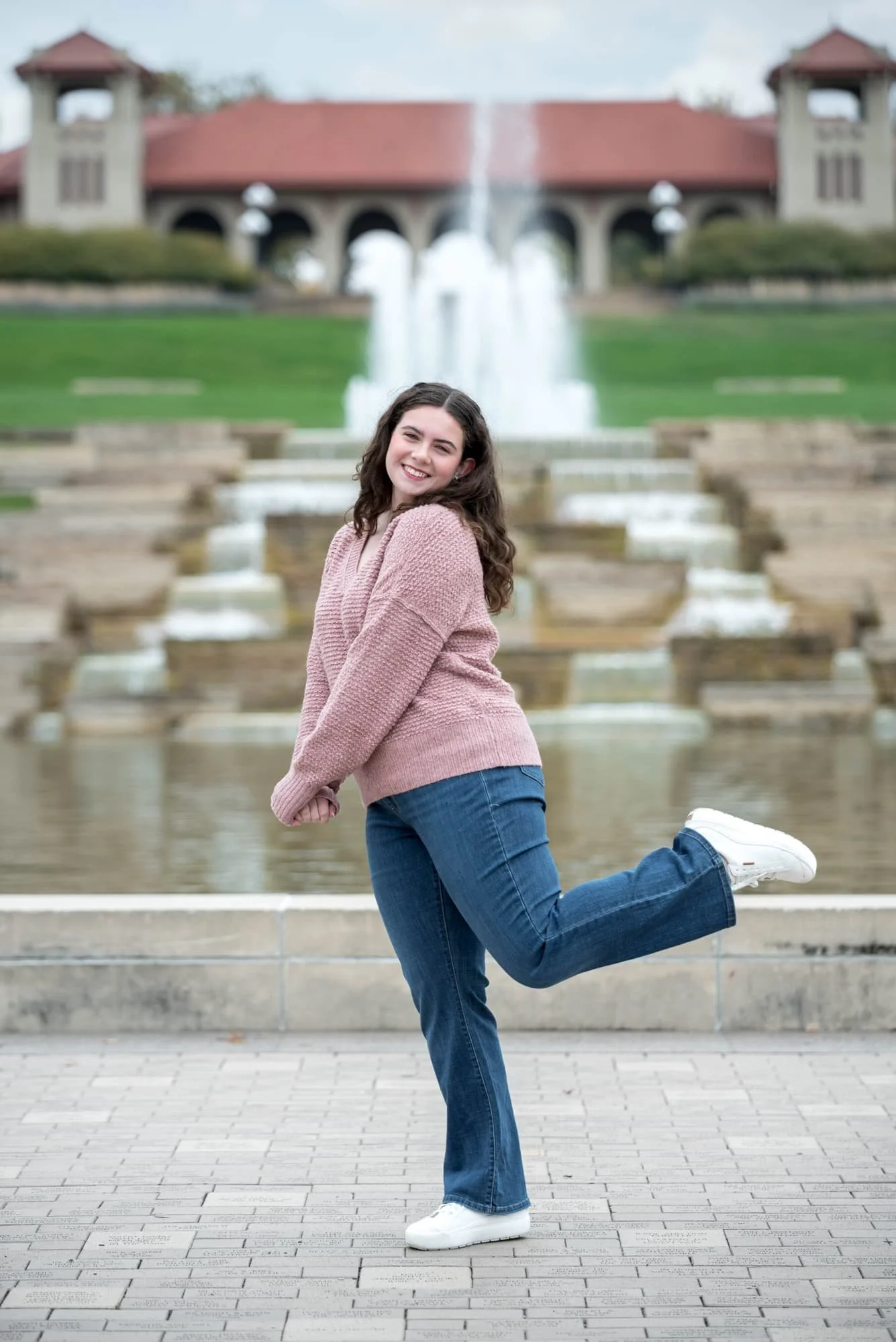 Standing outside the historic World's Fair Pavilion waterfall, a senior girl wearing a pink sweater and blue jeans smiles for a photo.