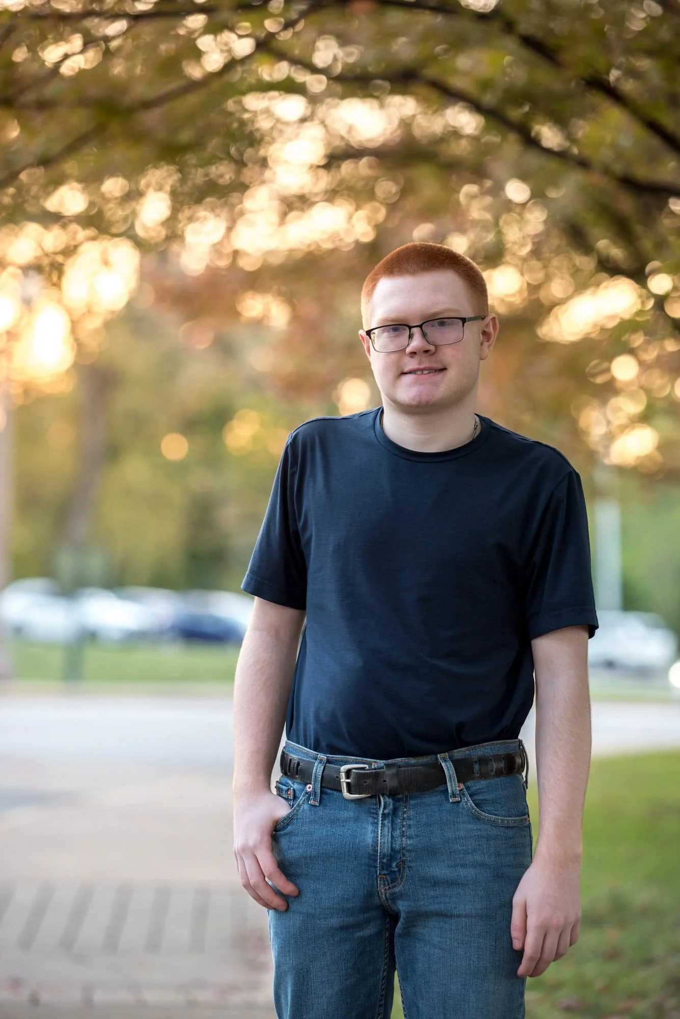 Sunlit nature photo showing a senior wearing a black t-shirt and blue jeans on a walking path as sunlight peeks through autumn trees at The Muny in Forest Park.