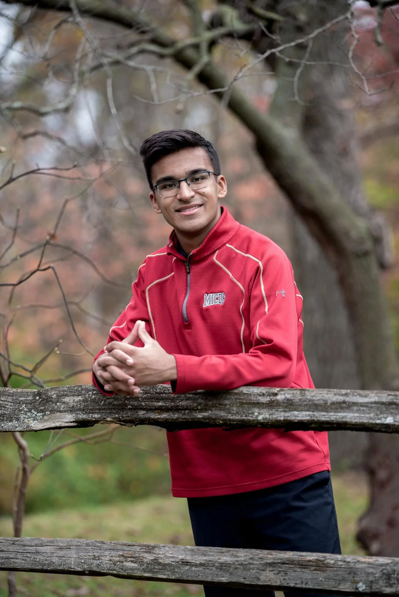Casual senior portrait of a guy in a red zip-up school shirt, leaning on a rustic wooden fence in front of a canopy of colorful fall trees in Chesterfield MO.