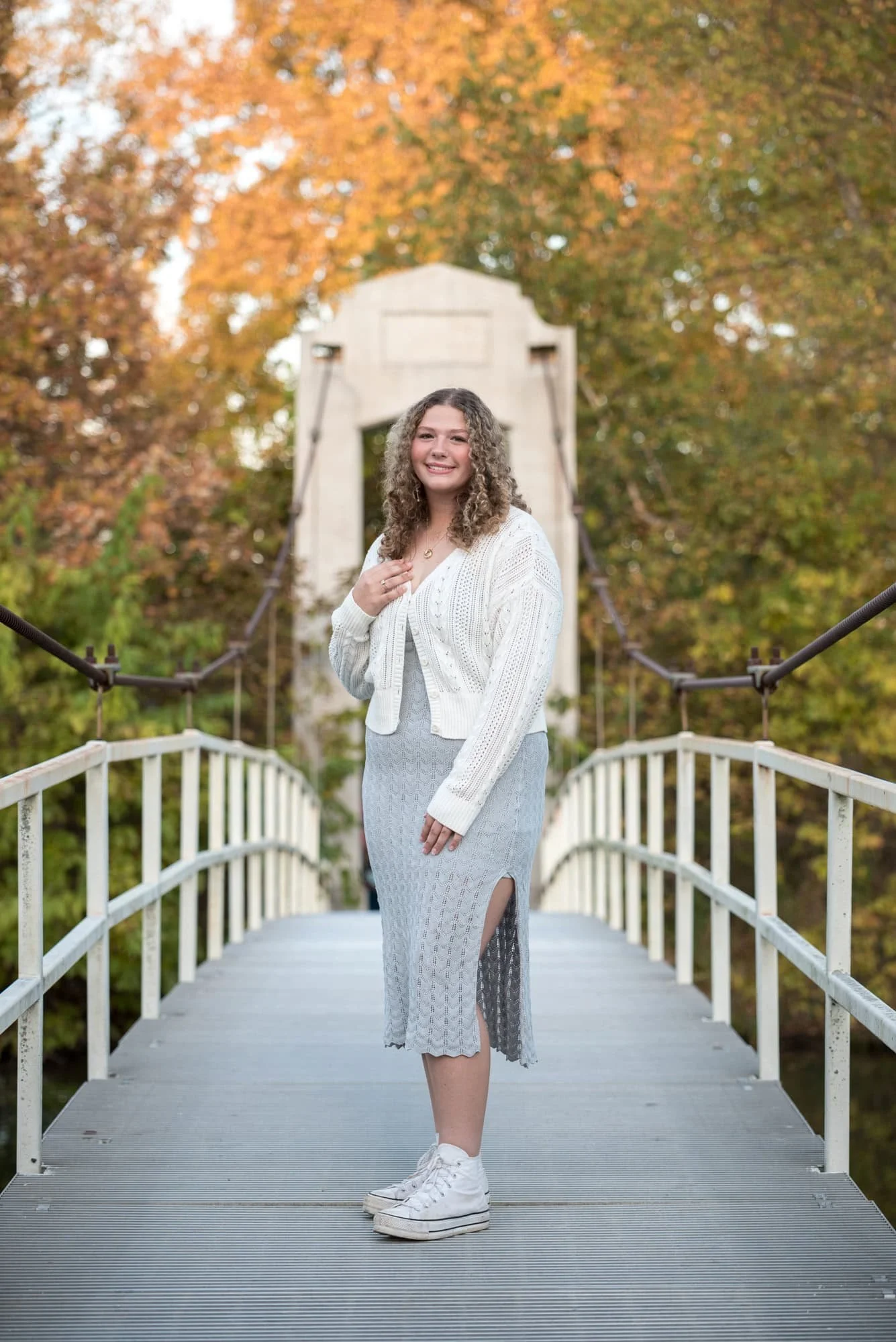 A St. Louis senior standing on a white suspension bridge near Art Hill wearing a white cardigan and gray maxi dress posing amidst peak fall colors in the region.