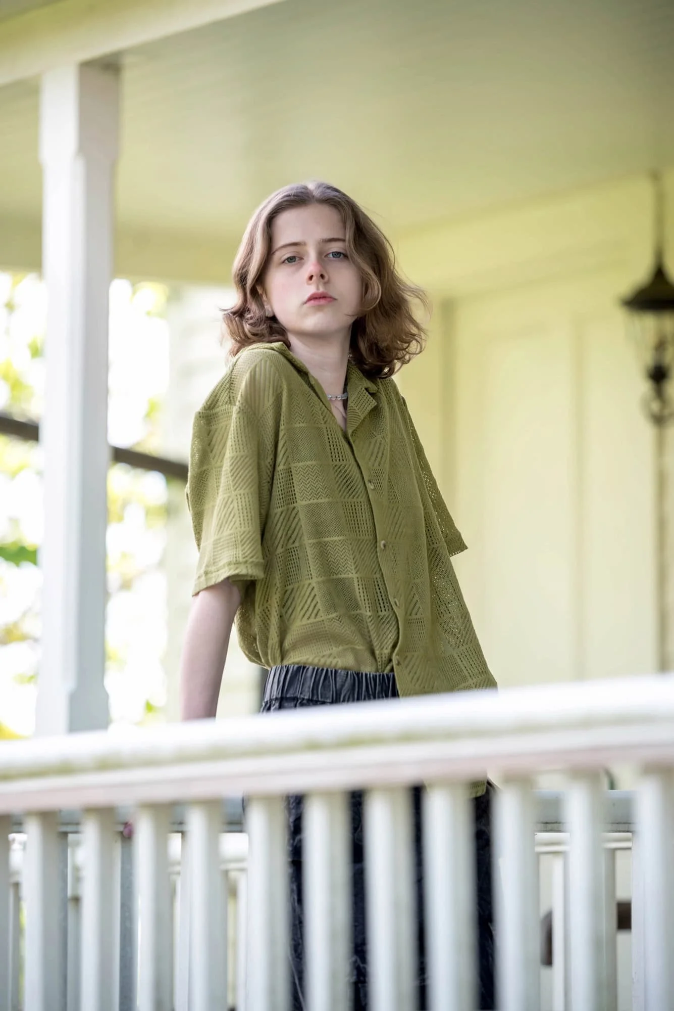 Low-angle senior photo of a girl looking down at the camera while leaning against a white wooden porch railing at a historic home at Shaw Nature Reserve, wearing an olive green button-up shirt and dark pants.