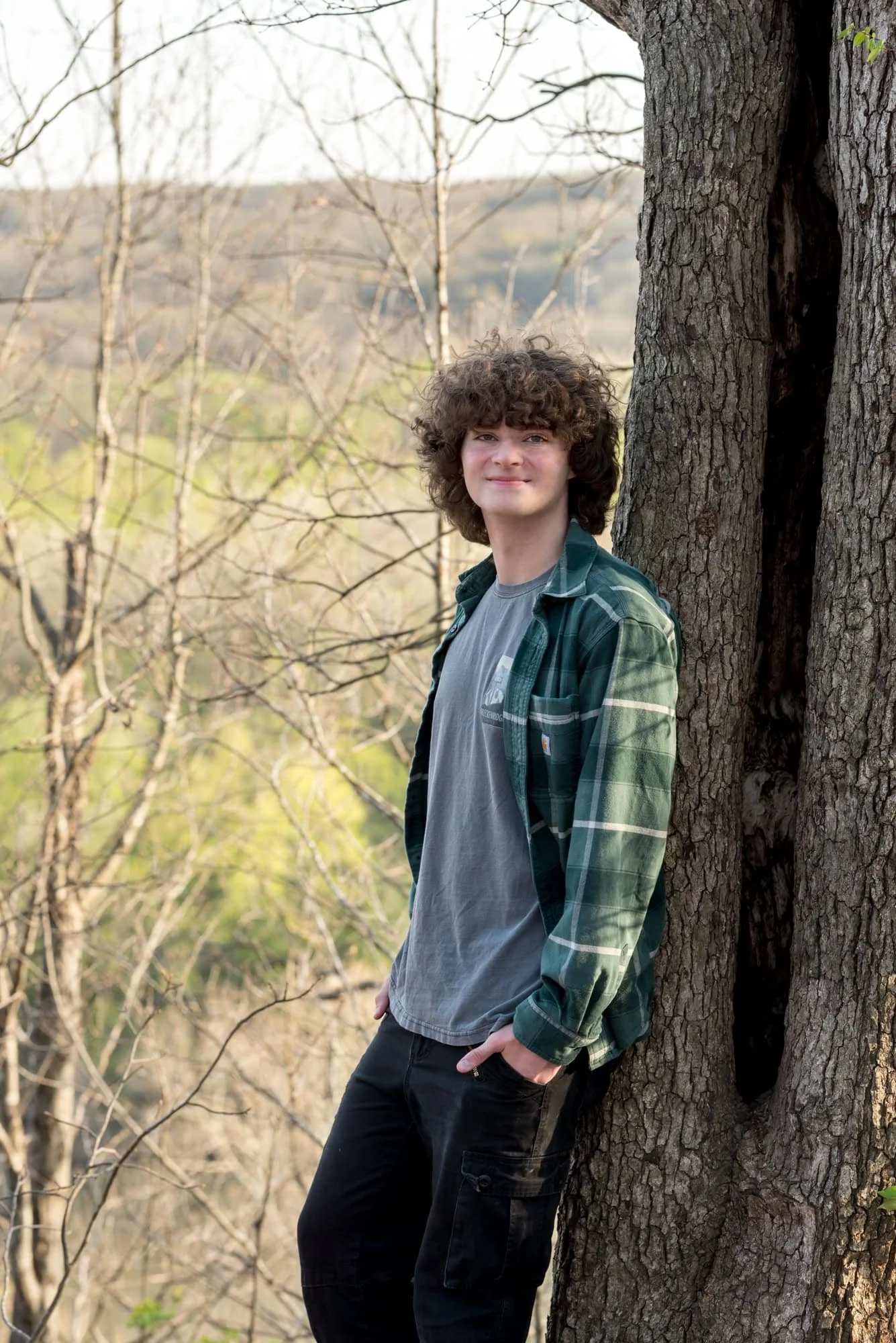 Senior photo of a guy smiling and leaning against a tree during a glowing spring sunset over the high overlook bluff at Castlewood State Park.