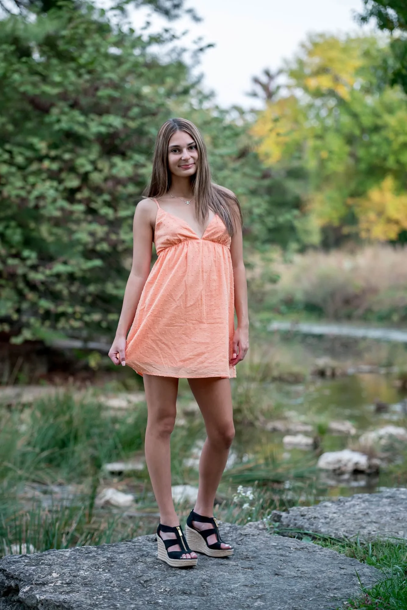 Classy high school senior picture featuring a girl in a short orange dress and black wedge heels near a creek by The Muny.