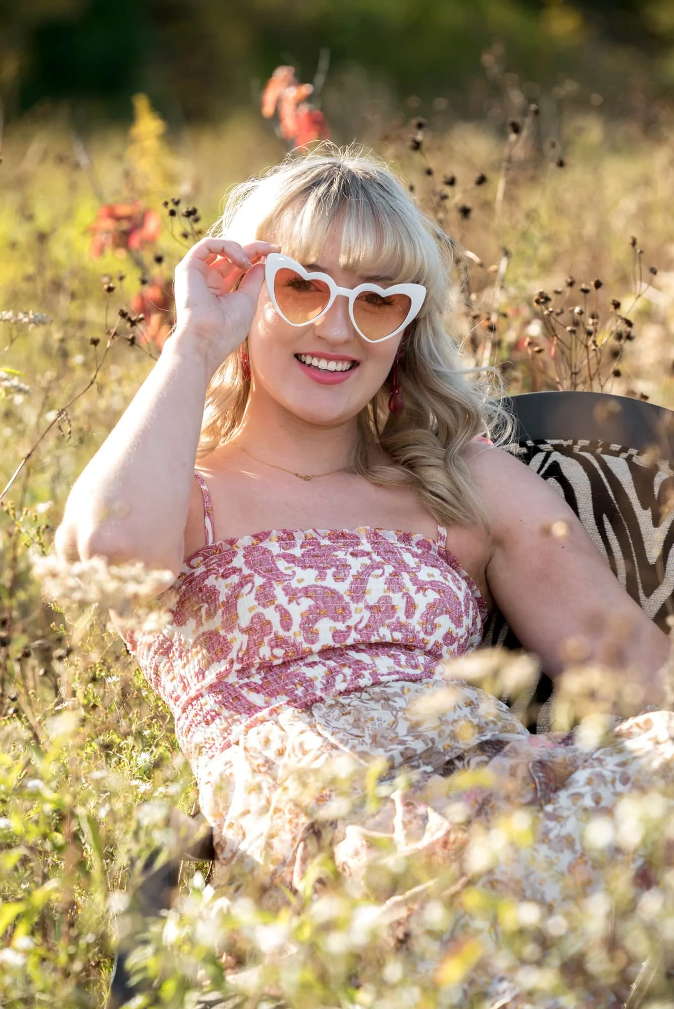 A senior girl wearing a pink and white floral dress sits on a chair in the tall grass for a picture taken by a Columbia, IL photographer.
