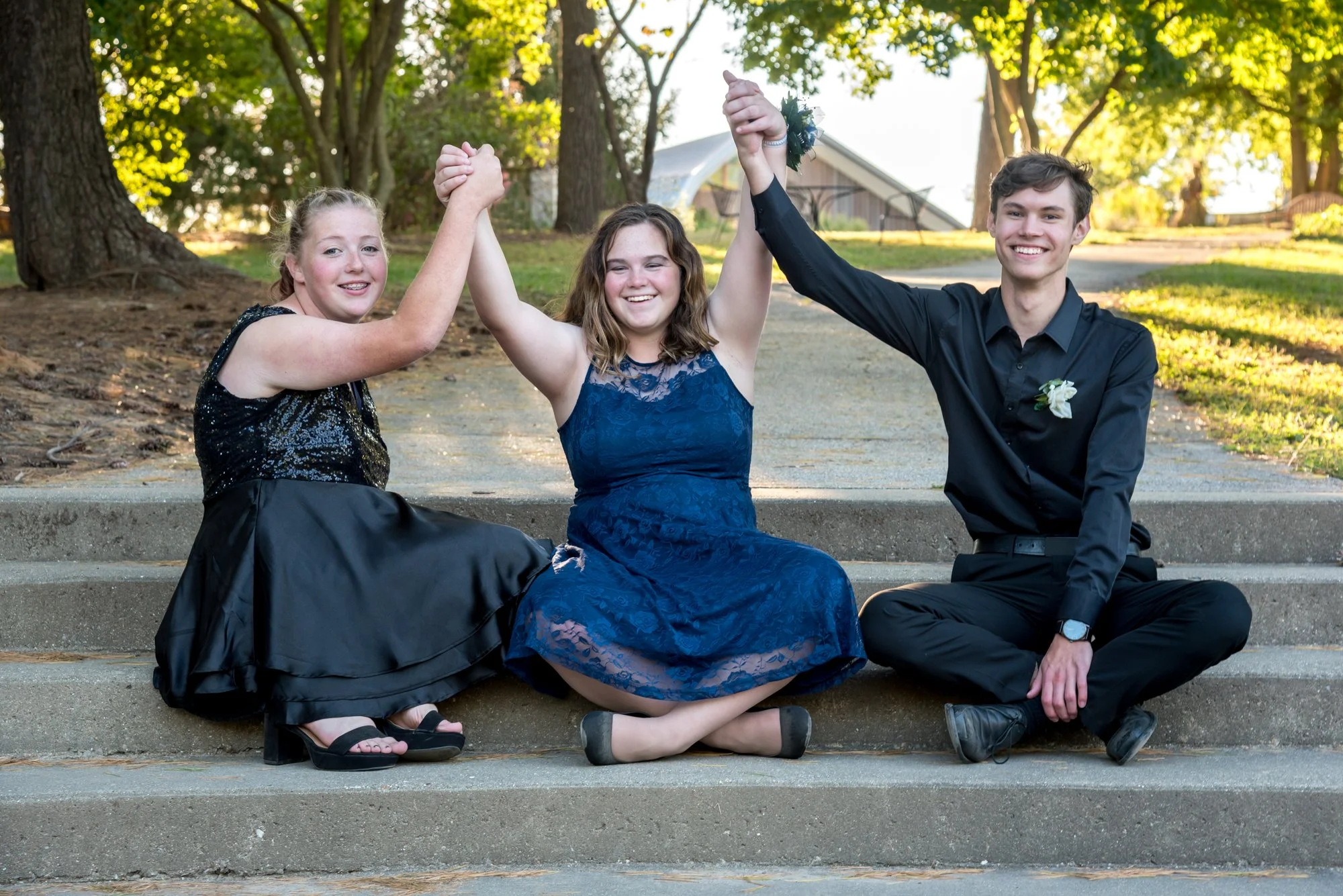 Joyful group homecoming picture at Laumeier Sculpture Park of three friends sitting on outdoor steps, holding hands and raising them high in the air as bright sunlight filters through the park trees behind them.