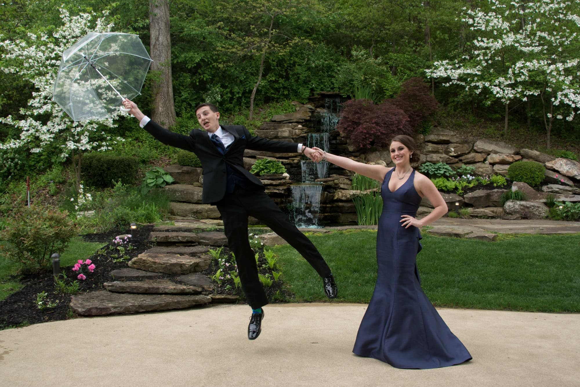 Action photo of a boy in a tuxedo playfully jumping into the air with a clear umbrella while holding his date's hand outside a venue in Monroe County, Illinois.