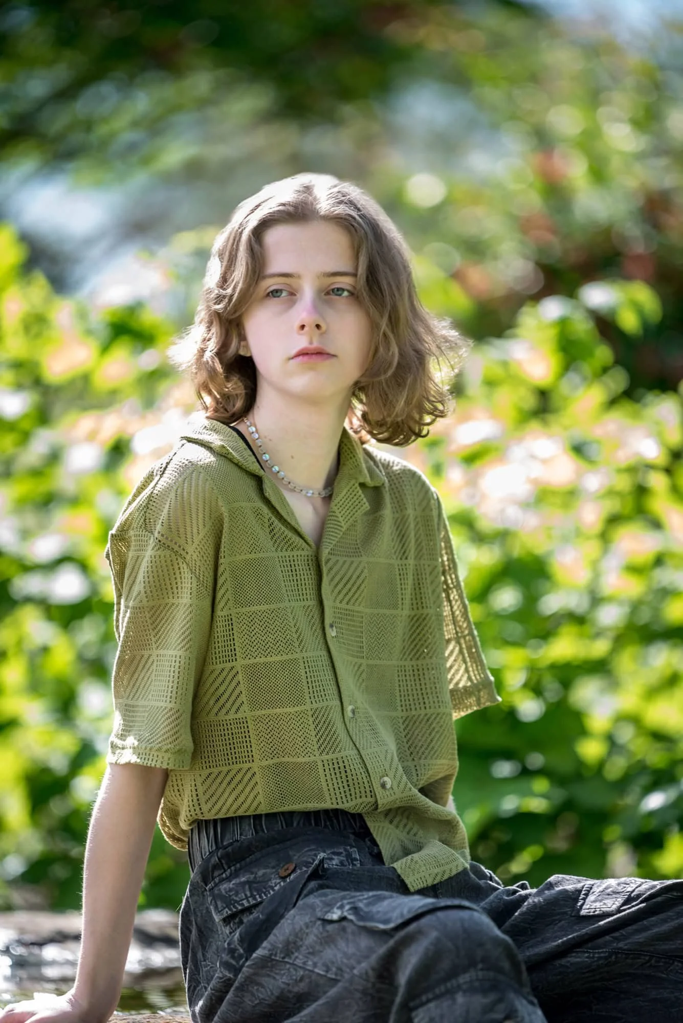 A vibrant outdoor senior picture from Franklin County showing a girl with a thoughtful look in natural light, sitting surrounded by the bright green foliage of sunlit summer bushes, wearing a textured olive-green shirt.