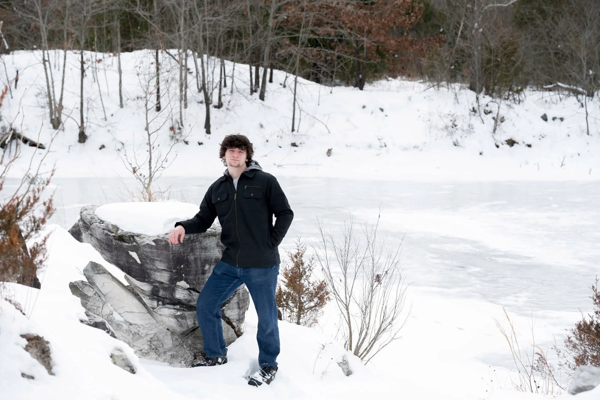 Crisp winter picture of a boy leaning against a large snow-covered boulder overlooking a frozen lake, work by a local Augusta Missouri senior photographer, wearing a black utility jacket and blue jeans.