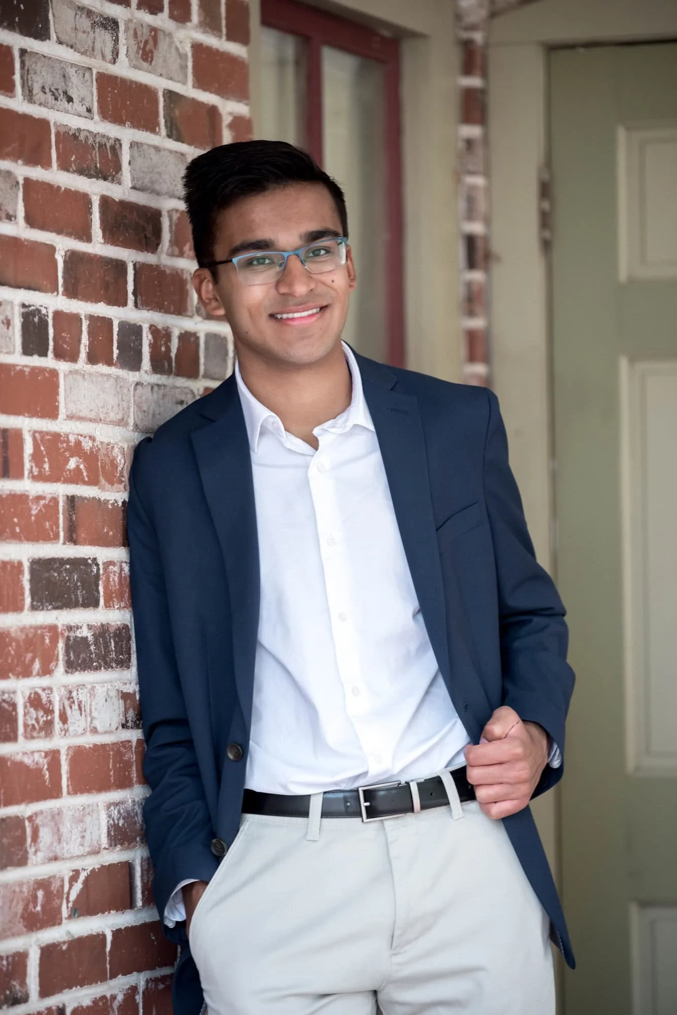 Sharp senior portrait of an Indian guy wearing a navy blue blazer and khakis, leaning against a classic brick building at Faust Park.