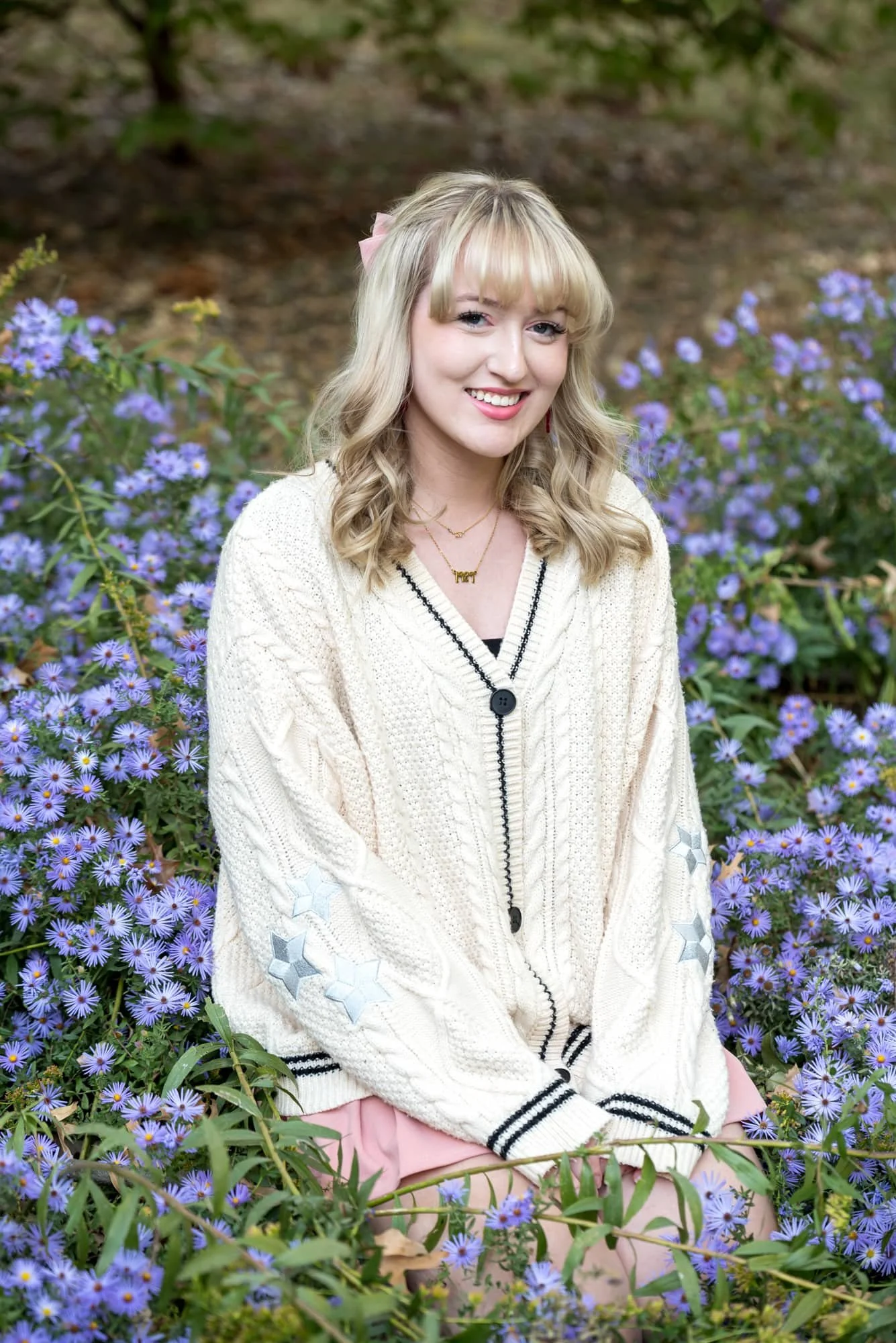 A high school senior girl wearing a white sweater and a pink skirt Taylor Swift inspired outfit sits in purple flowers at Lafayette Park in St. Louis.