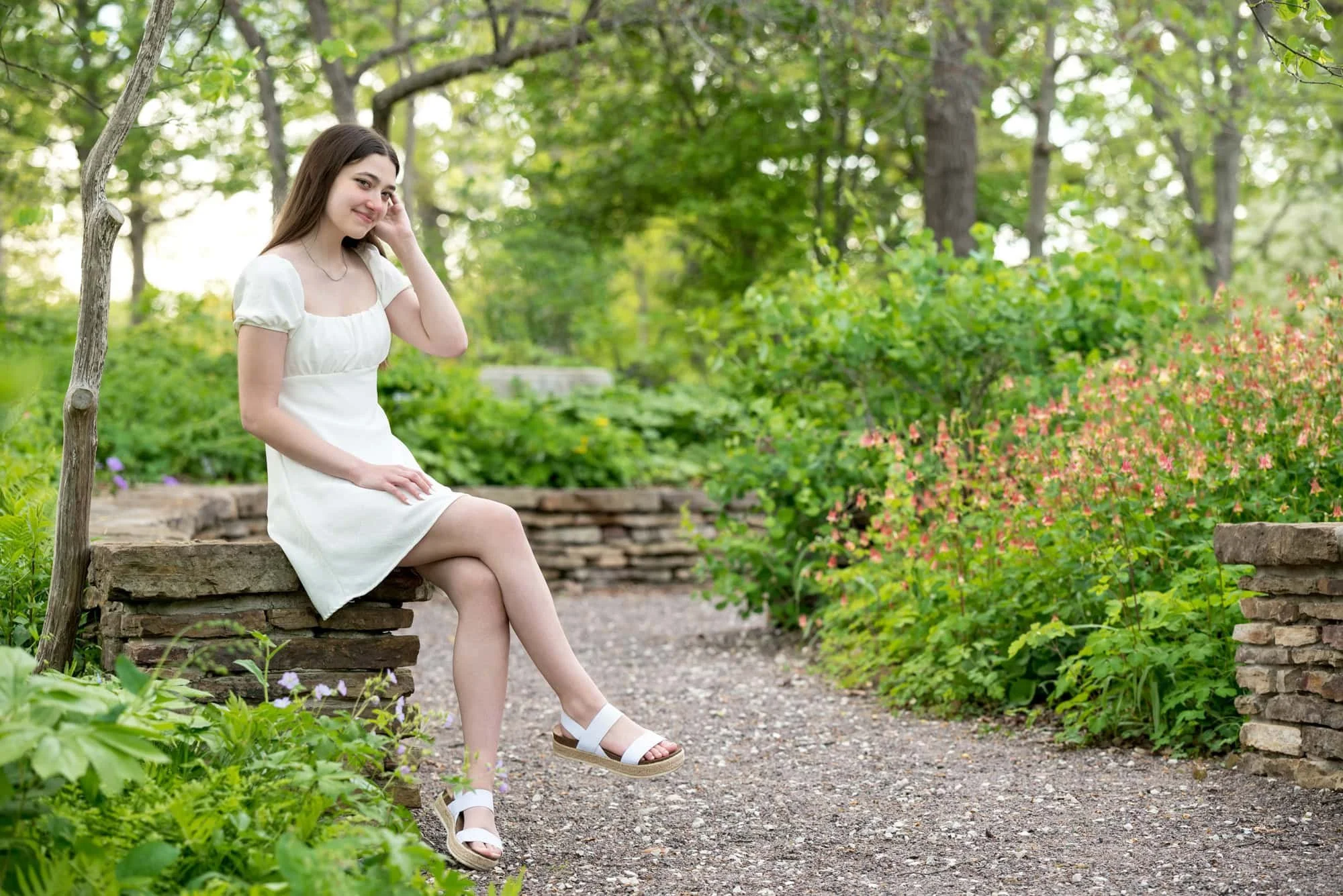 Graduating senior girl wearing a short white dress and white sandals, sitting gracefully on a historic stone wall surrounded by lush flowers and mature trees at Shaw Nature Reserve in Gray Summit MO.