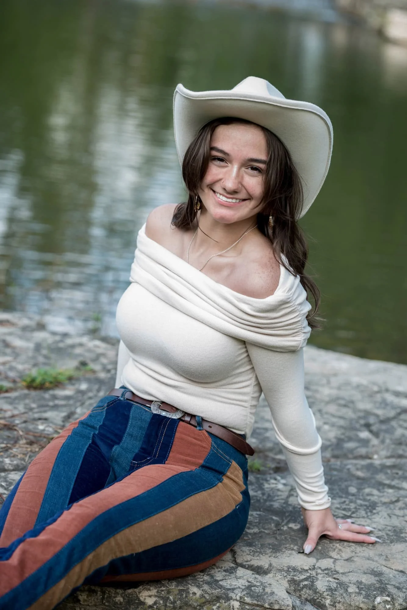Sitting on a large rock near a creek, taken by a St. Louis photographer, a senior girl in a white cowboy hat and striped blue jeans poses for a photo.