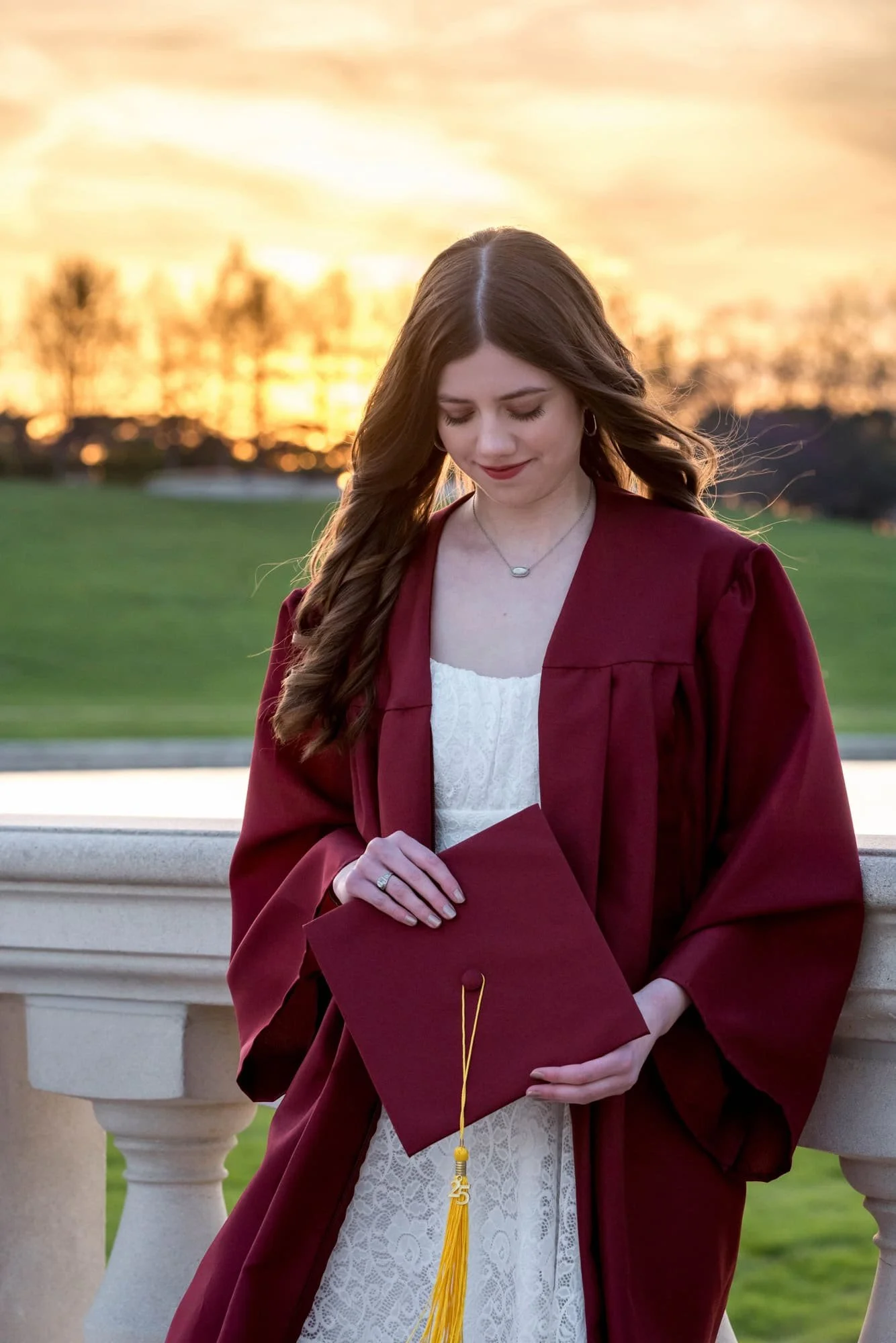 High school graduate wearing a short white dress and her maroon cap and gown, standing against a bridge at the Art Hill lake basin during a vibrant orange sunset, looking down at her cap with a soft smile.