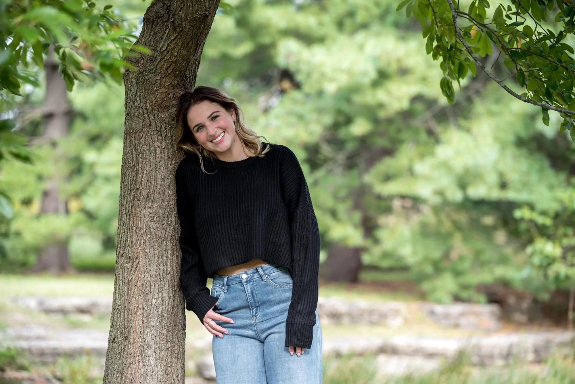 Natural rustic scene capturing a girl wearing a black sweater and blue jeans while leaning against a large tree trunk taken by a St. Louis senior photographer.