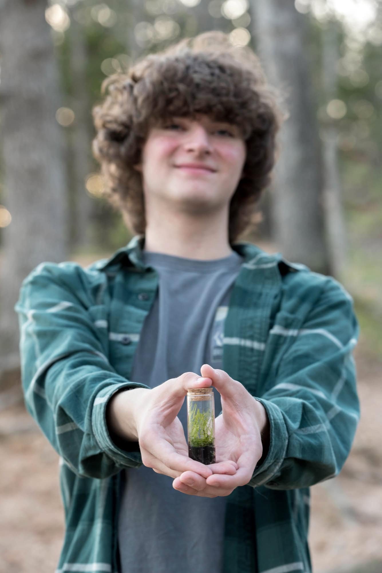 Senior guy smiling and holding out a small terrarium while standing in the middle of the woods, wearing a green plaid shirt for a creative portrait at Castlewood State Park in Ballwin.
