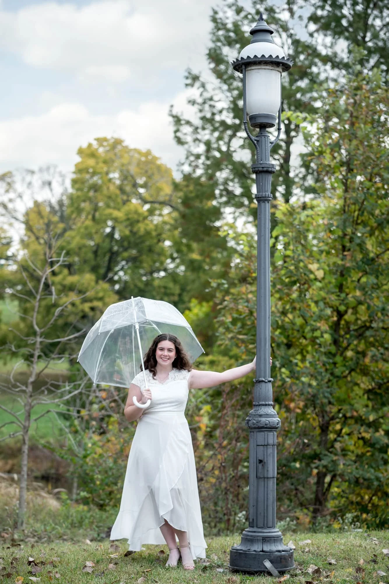 Standing next to a park lamp while holding an umbrella, a senior girl in a white dress poses for an outdoor photograph in Forest Park.