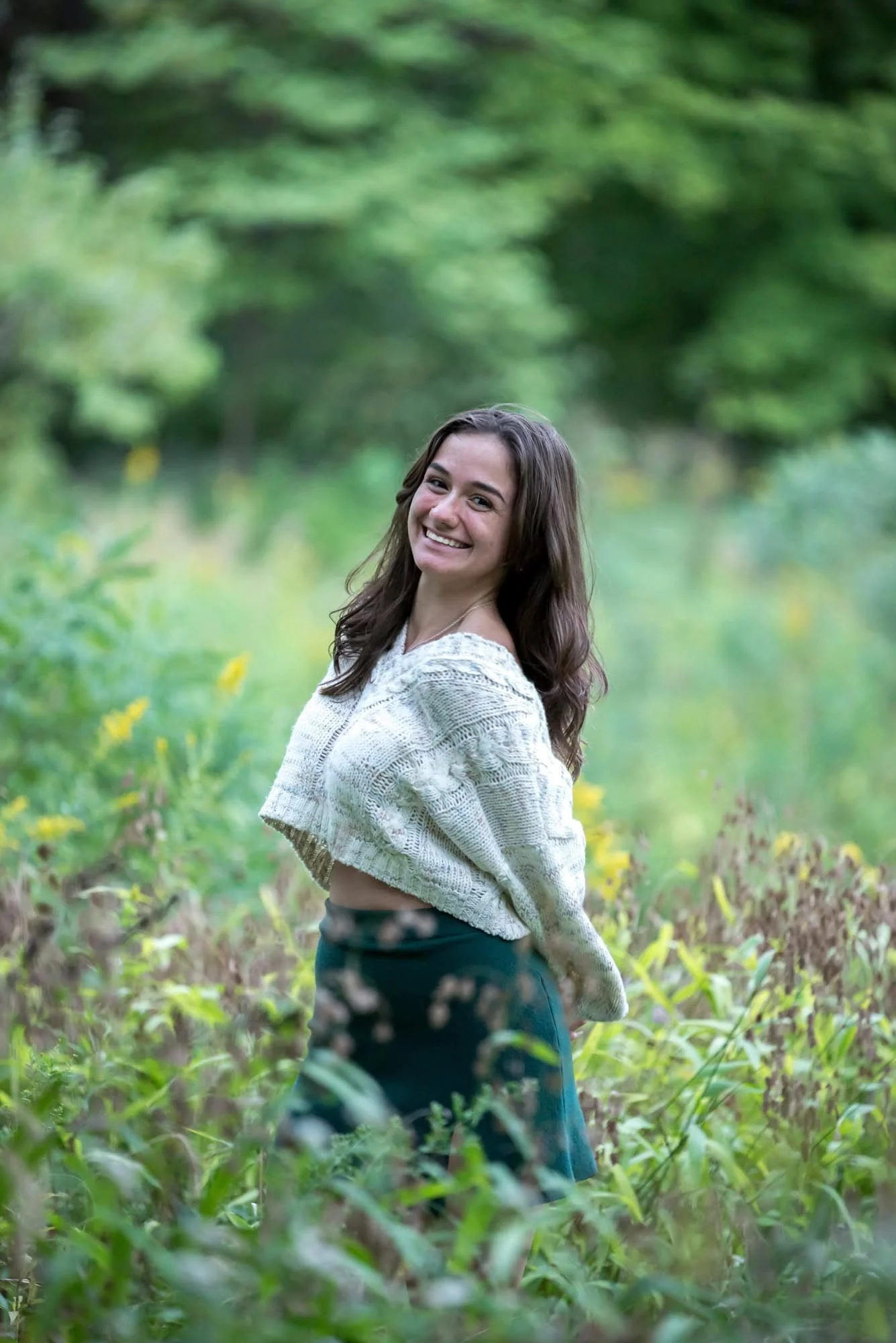 Posing in a field of tall grasses in St. Louis County, a senior girl wearing a white sweater and green skirt smiles for a picture.