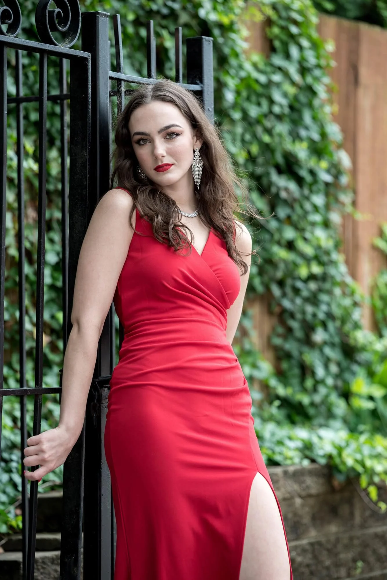 A photo captures a high school senior girl wearing a red long dress as she leans against a wrought iron fence covered in green ivy in The Loop.