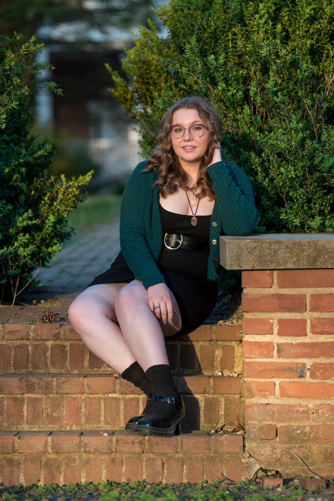 Sitting outdoors on brick steps during sunset in Town and Country, a senior girl in a black dress and green cardigan smiles for a picture.