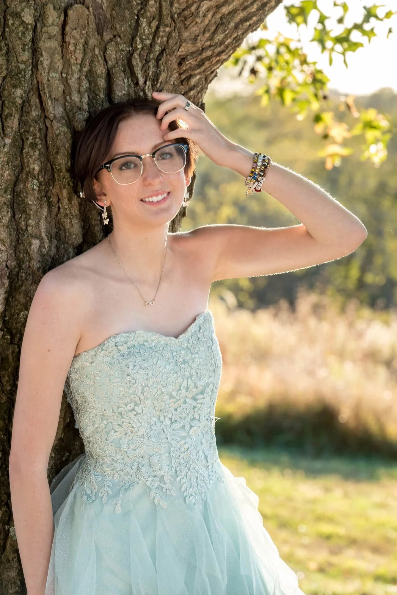 Close-up portrait senior photo of a girl with her hand in her hair, wearing her green prom dress while leaning against a rustic tree trunk with tall grasses blurred in the background at Queeny Park.