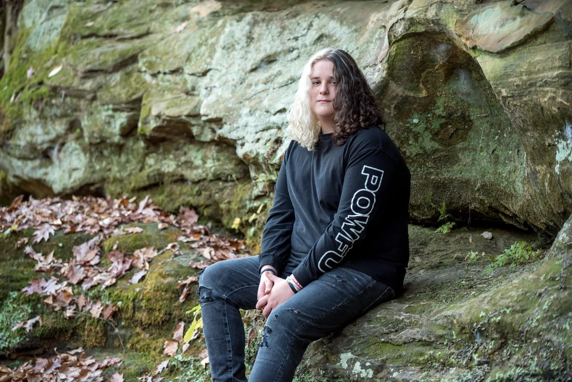 Natural autumn senior portrait of a boy with half-brown and half-white hair, sitting on a mossy rock amidst fallen leaves wearing a black sweatshirt and ripped black jeans in Monroe County.