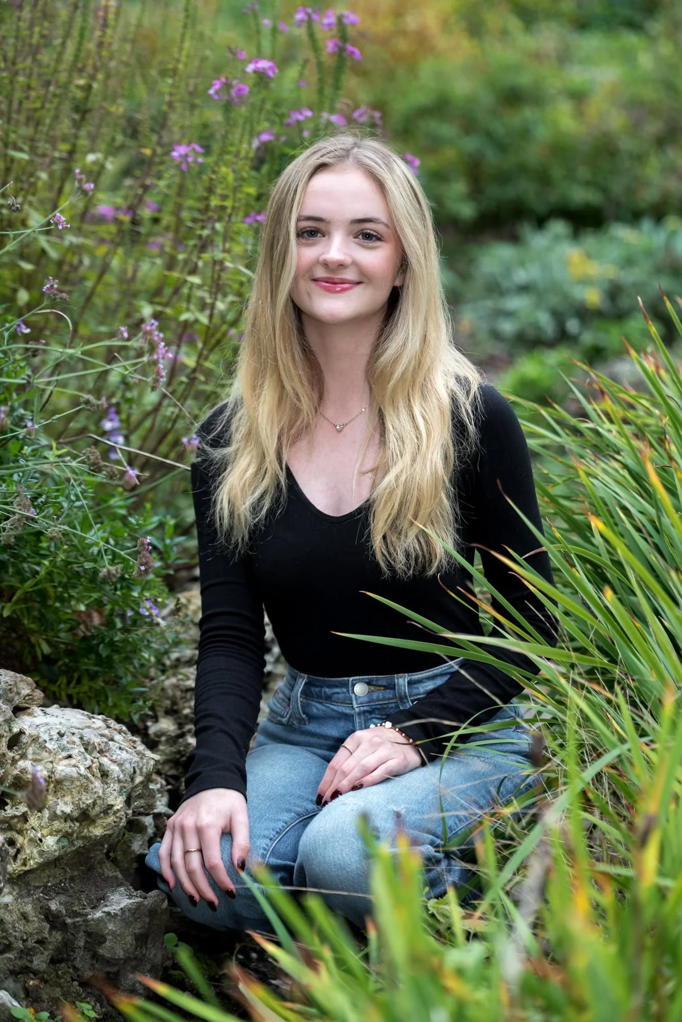 Sitting among seasonal fall flowers, a St. Louis senior girl in a casual black long-sleeve top and blue jeans smiles for a photo.