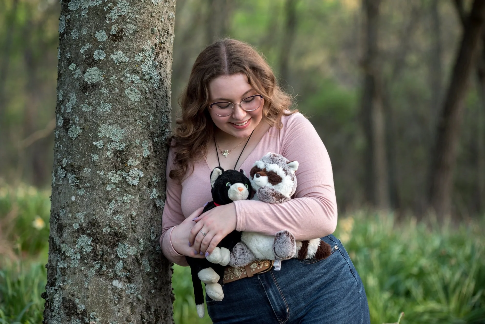 A photo shows a high school senior girl wearing a pink long-sleeve shirt and blue jeans at Queeny Park leaning on a tree and looking down at childhood stuffed animals.