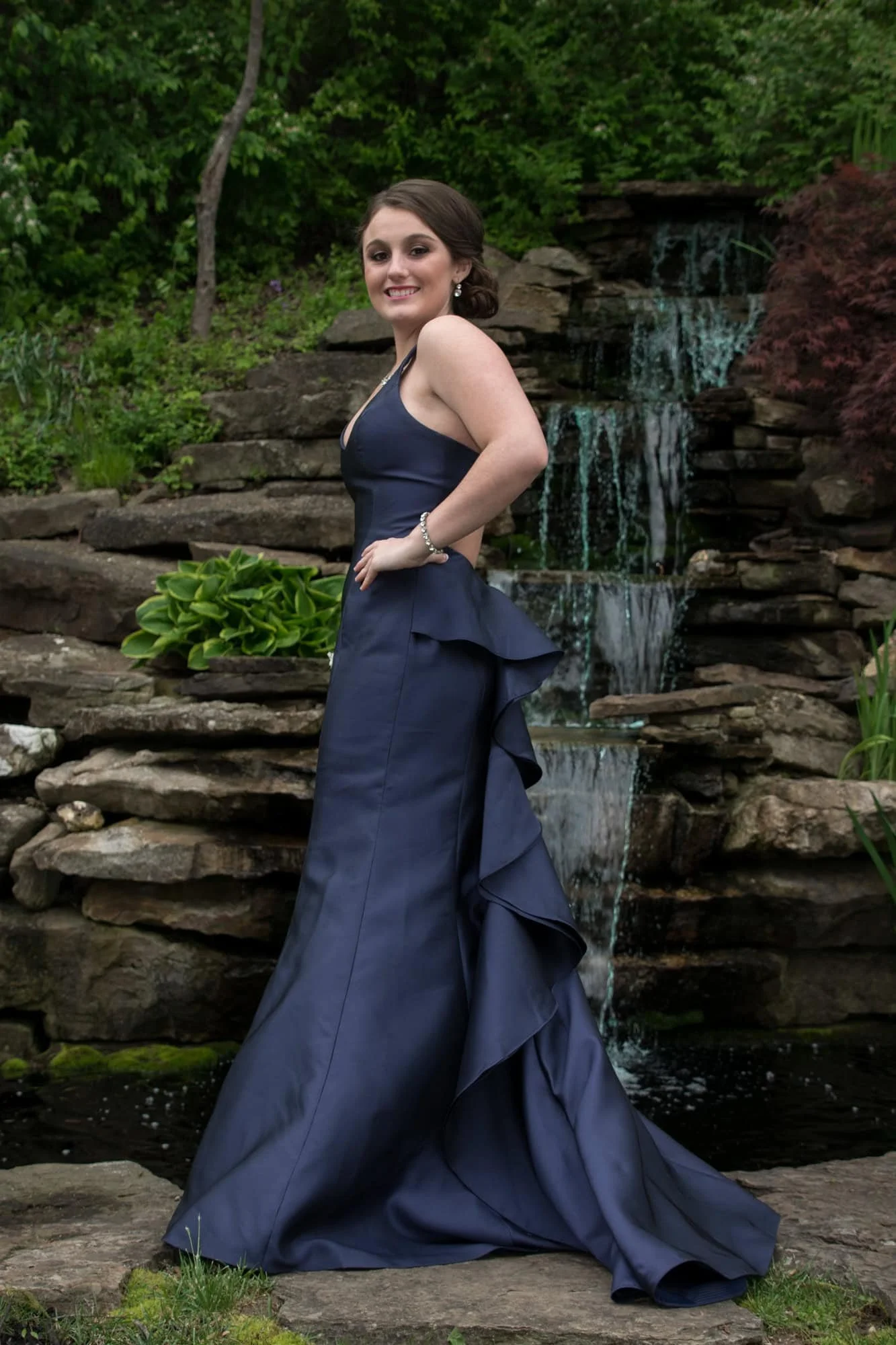 Full-length photo of a girl posing in her navy blue satin prom gown with a ruffled train, standing on stone steps in front of a tiering rock waterfall in Monroe County.