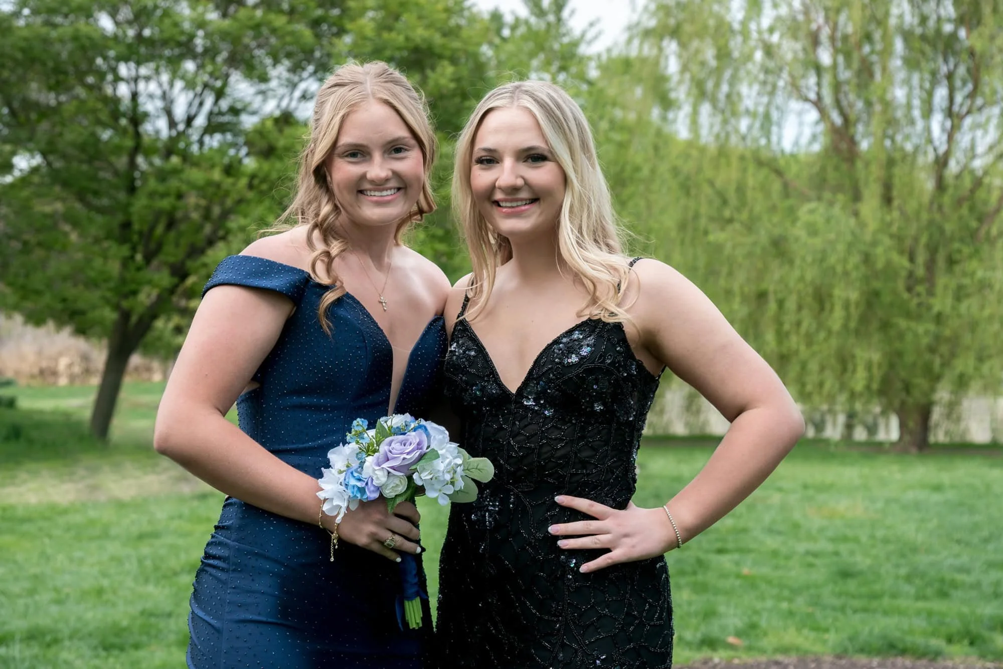 A picture captures a senior girl in a navy blue dress standing in a grassy area with a friend in a black prom dress at Tower Grove Park.