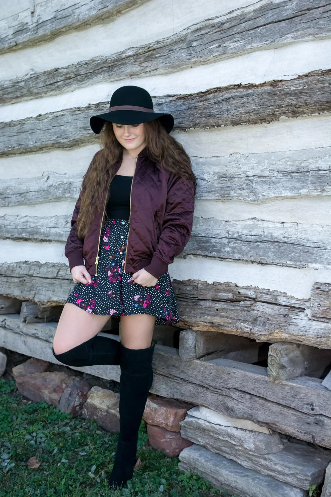 Edgy boho senior portrait by a Waterloo IL senior photographer, showing a girl wearing a black floppy hat, a purple jacket over a black floral dress, and knee-high black boots, leaning back against a rustic log cabin while looking down.