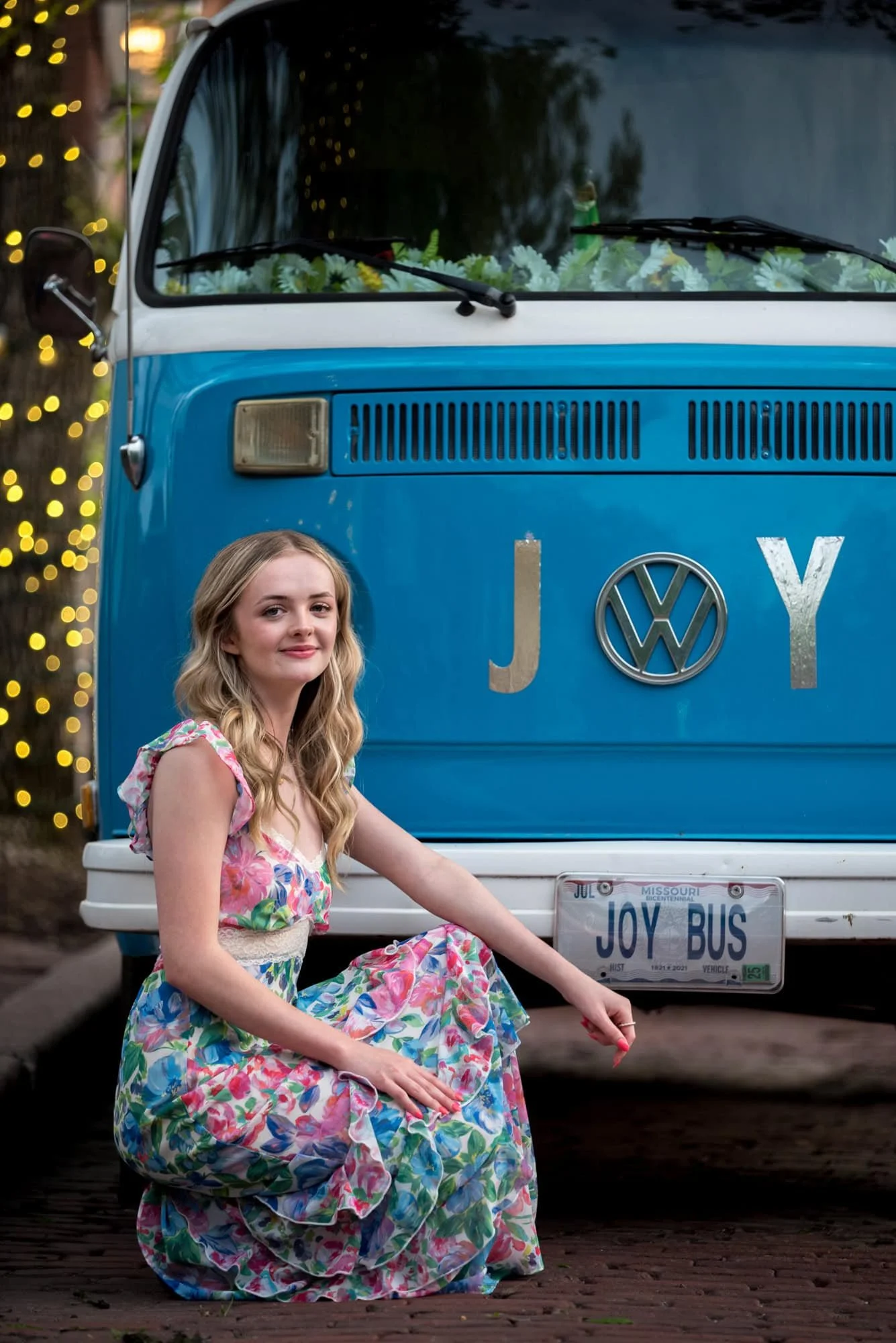 A spring photo, taken by St. Charles County photographer, shows a high school senior girl wearing a floral long dress sitting in front of a vintage Volkswagen bus.