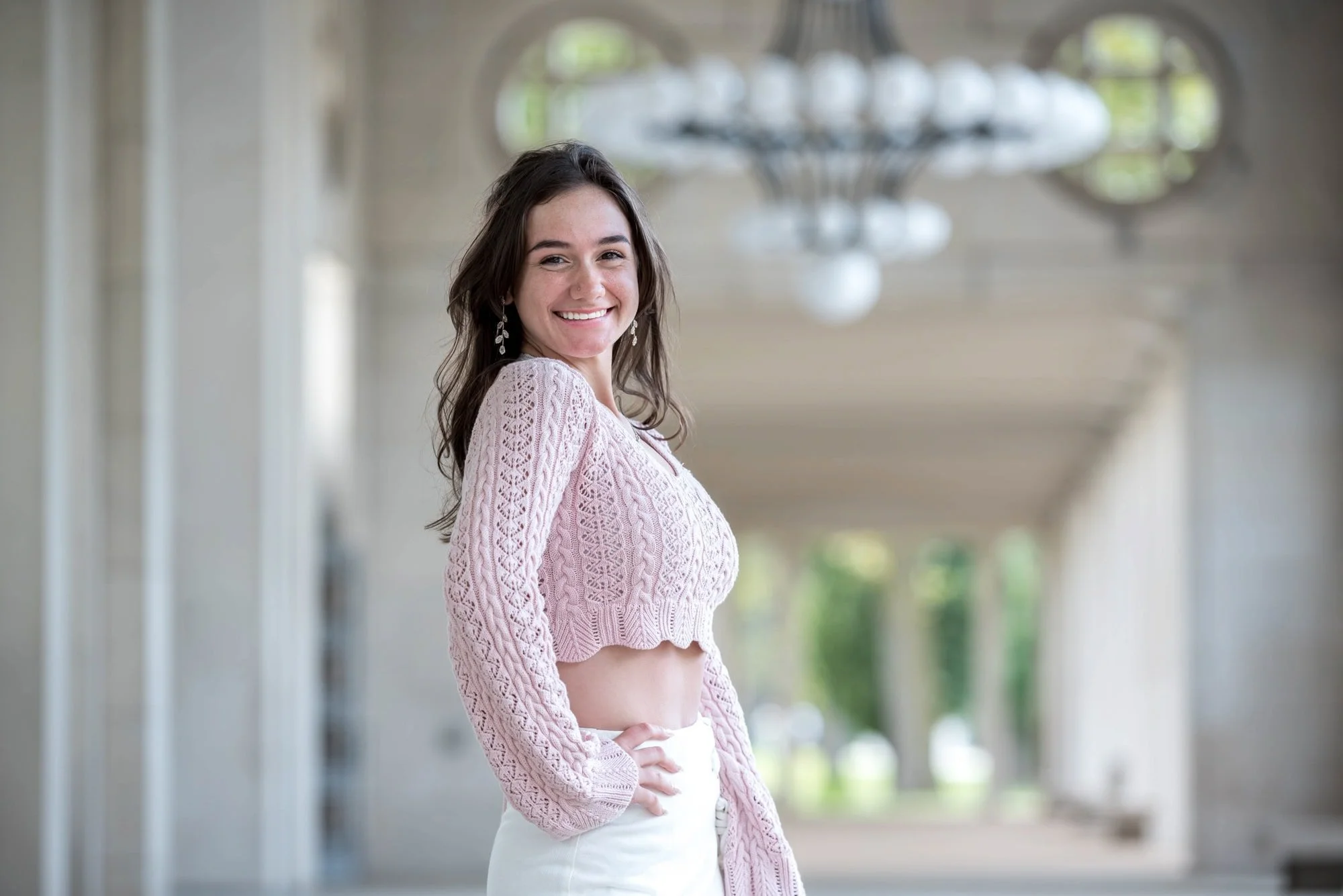 Chic and whimsical senior picture of a smiling girl wearing a pink knit cropped top and a white skirt, posing turned sideways with her hand on her hip directly underneath a grand historic chandelier at The Muny in Forest Park.