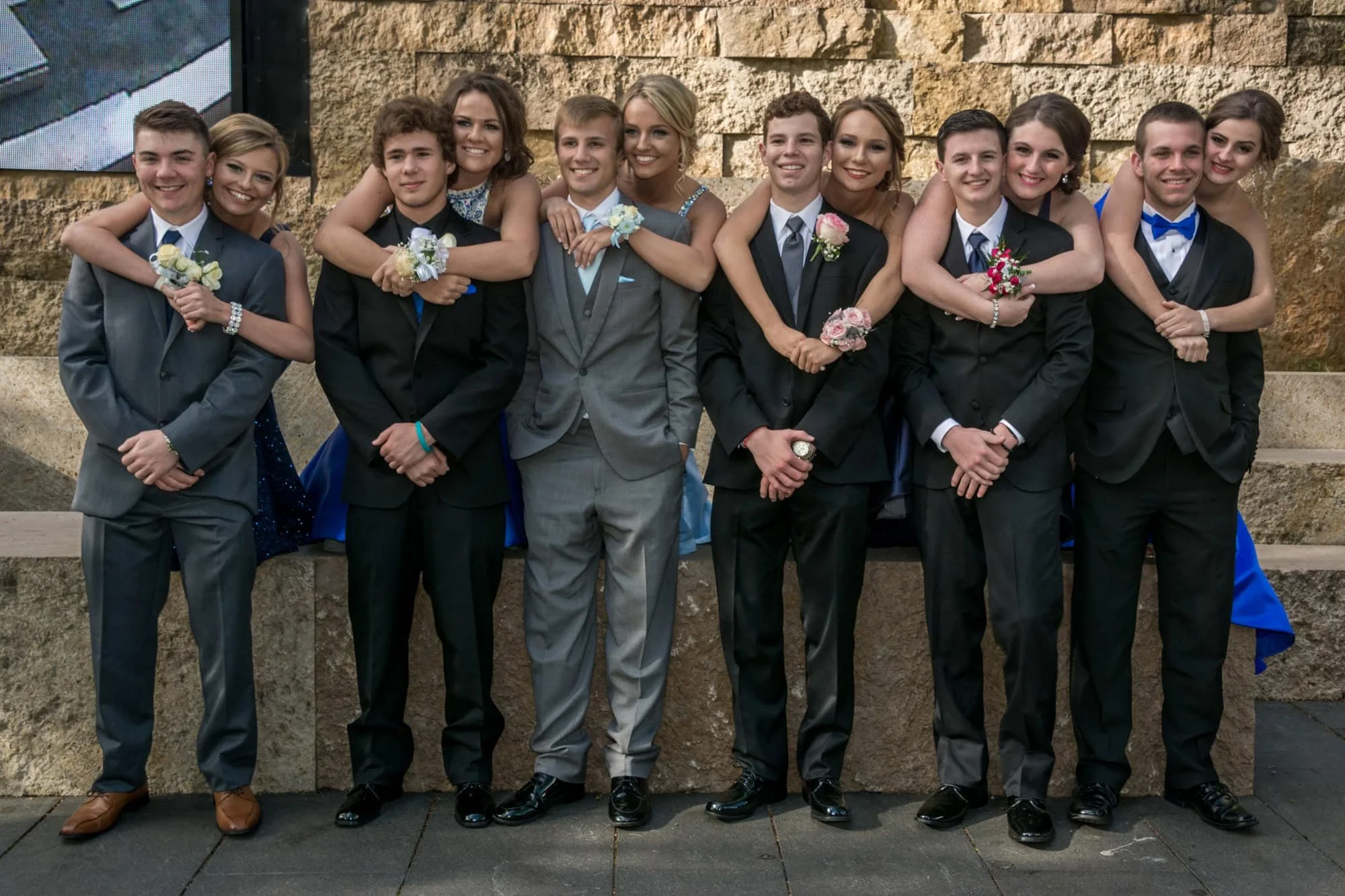 A line of six high school prom couples posing together with the girlfriends arms around their boyfriends against a stone block wall in downtown St. Louis.