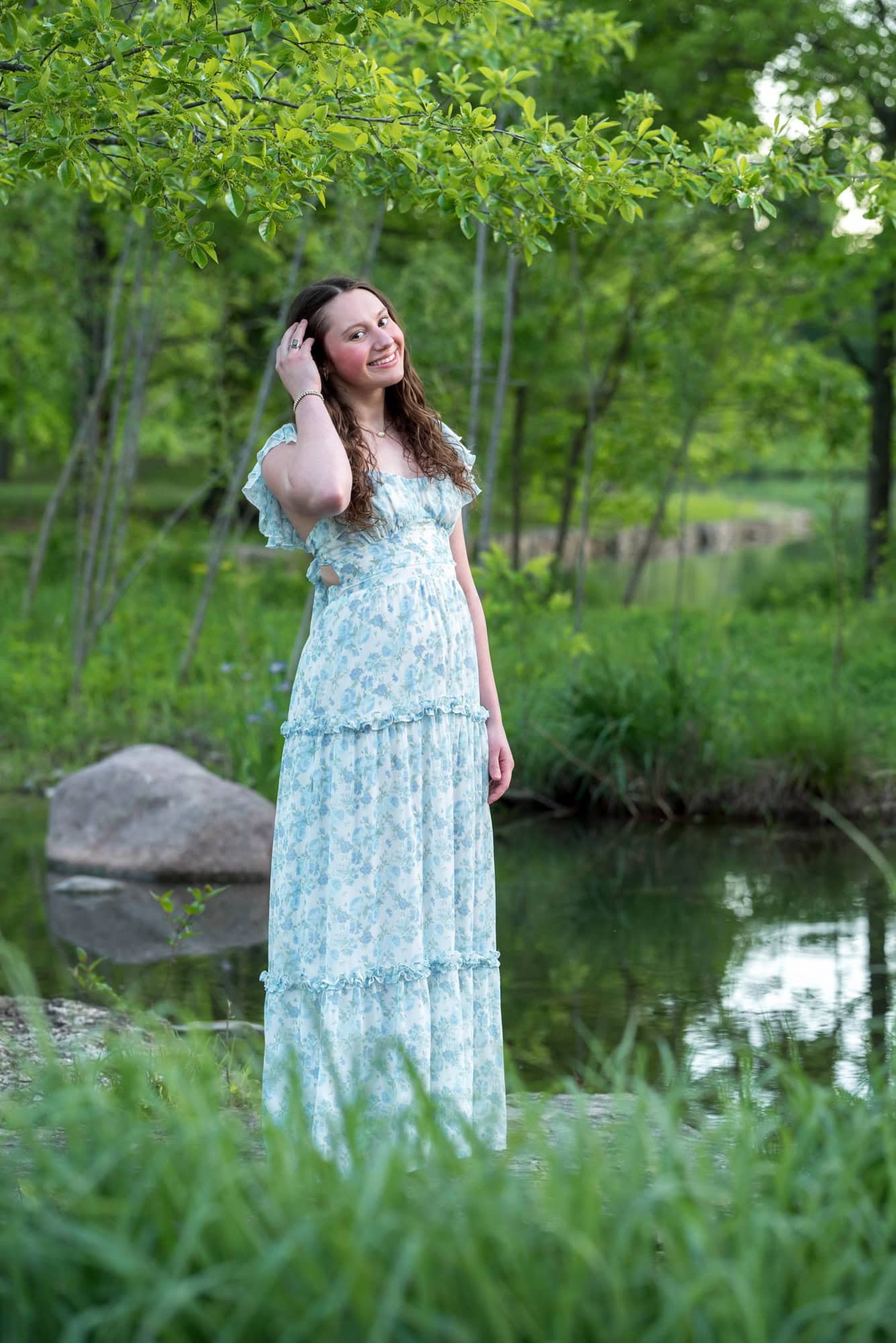Standing by a rocky creek near Art Hill, a high school senior girl smiles gently while fixing her long curly hair.