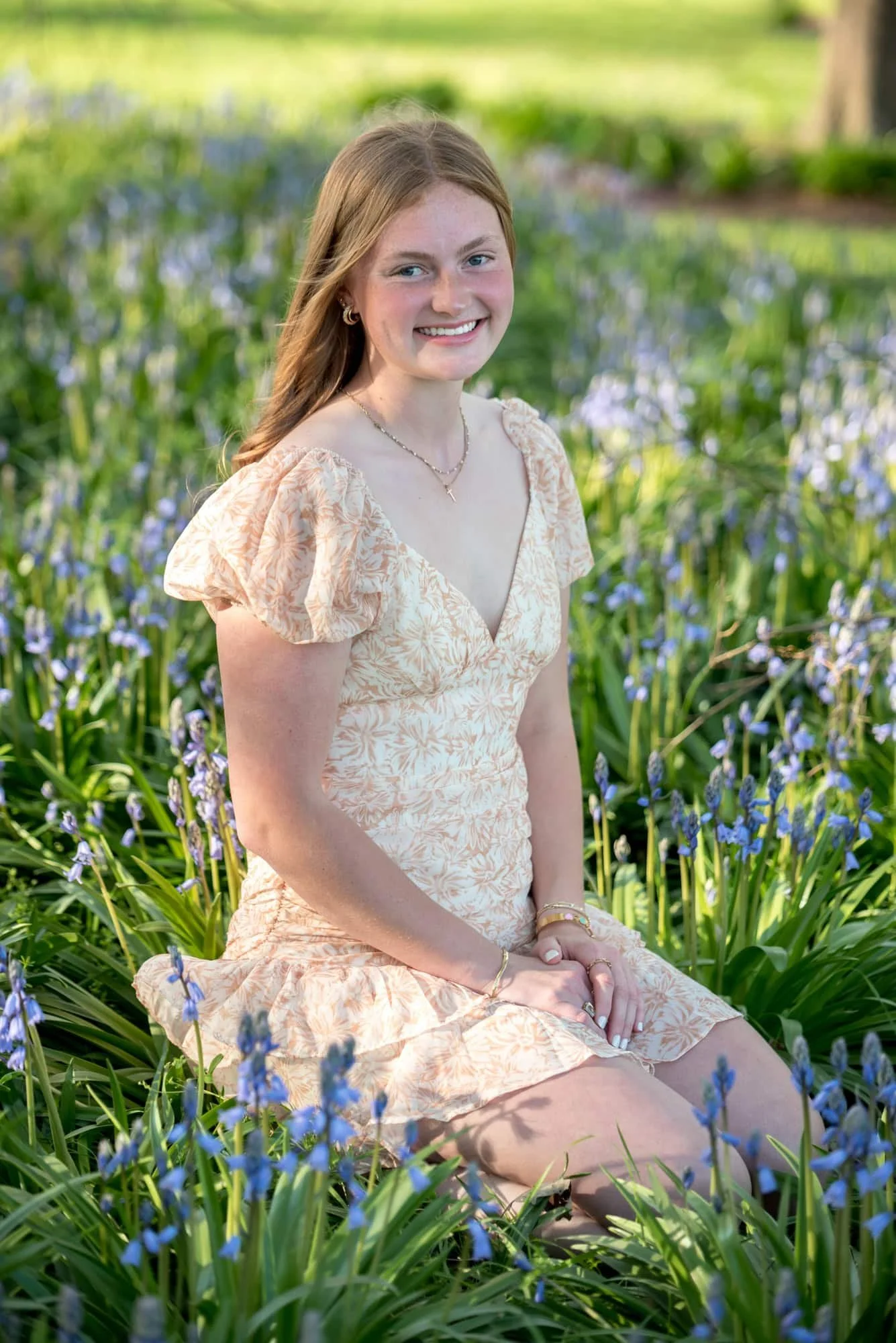 Sitting in a patch of purple spring flowers, a senior girl in an orange and white dress poses for a photo, taken by a St. Louis County photographer.