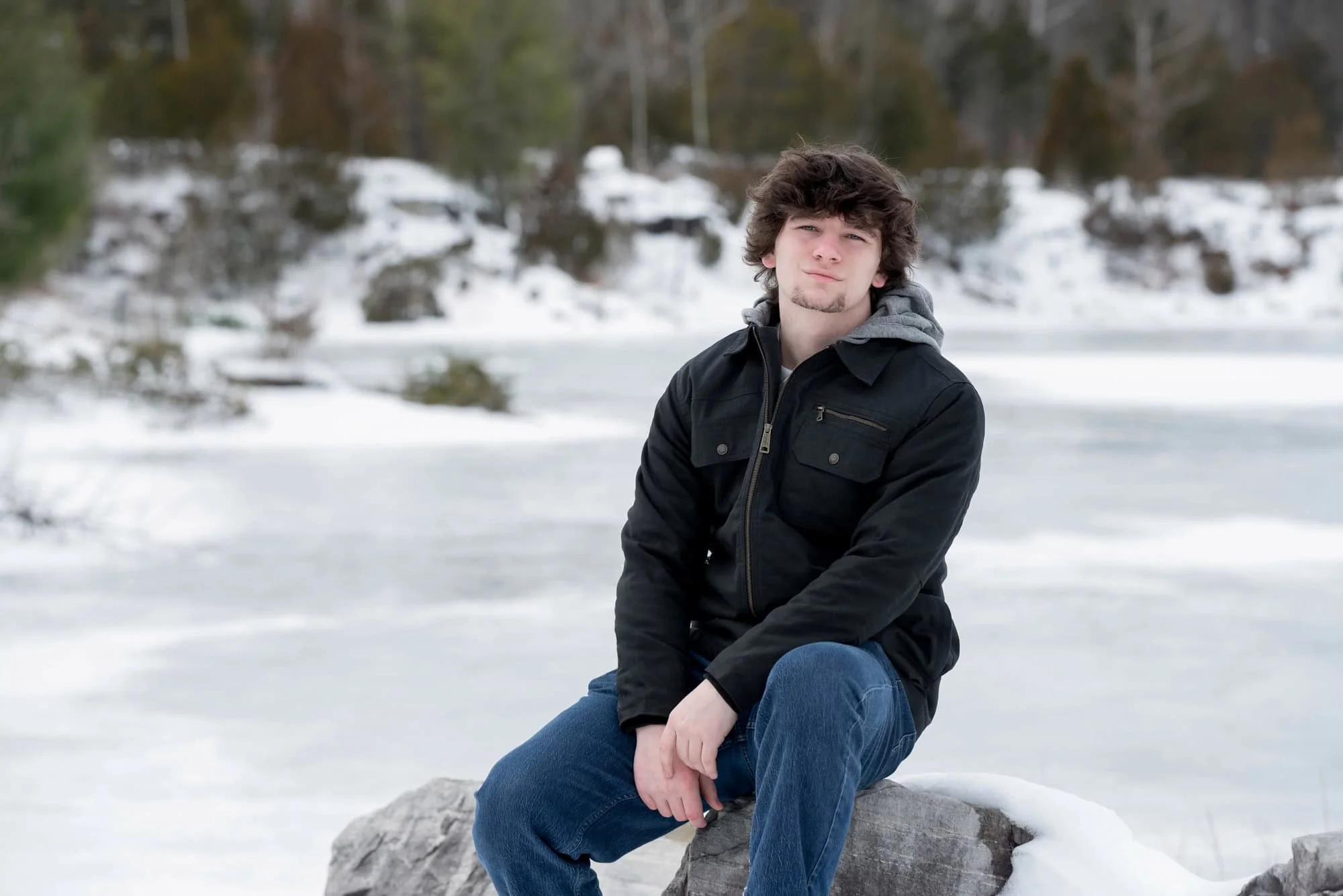 High school senior boy in a black winter jacket on a snow-covered rock with a frozen lake behind him at Klondike Park.