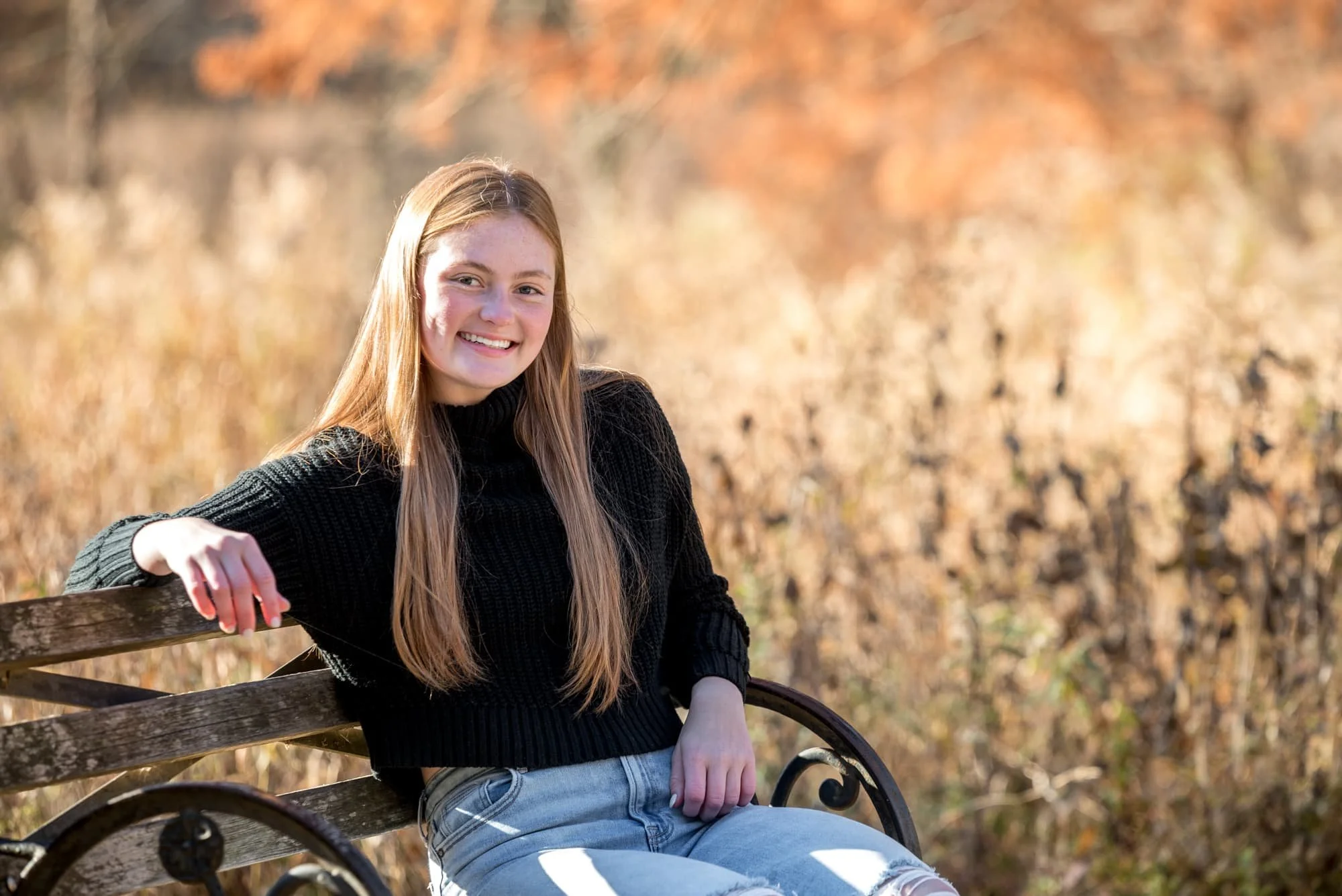 A morning golden hour photo captures a high school senior girl wearing a black sweater and blue jeans in St. Louis County sitting on a bench in front of tall grasses.
