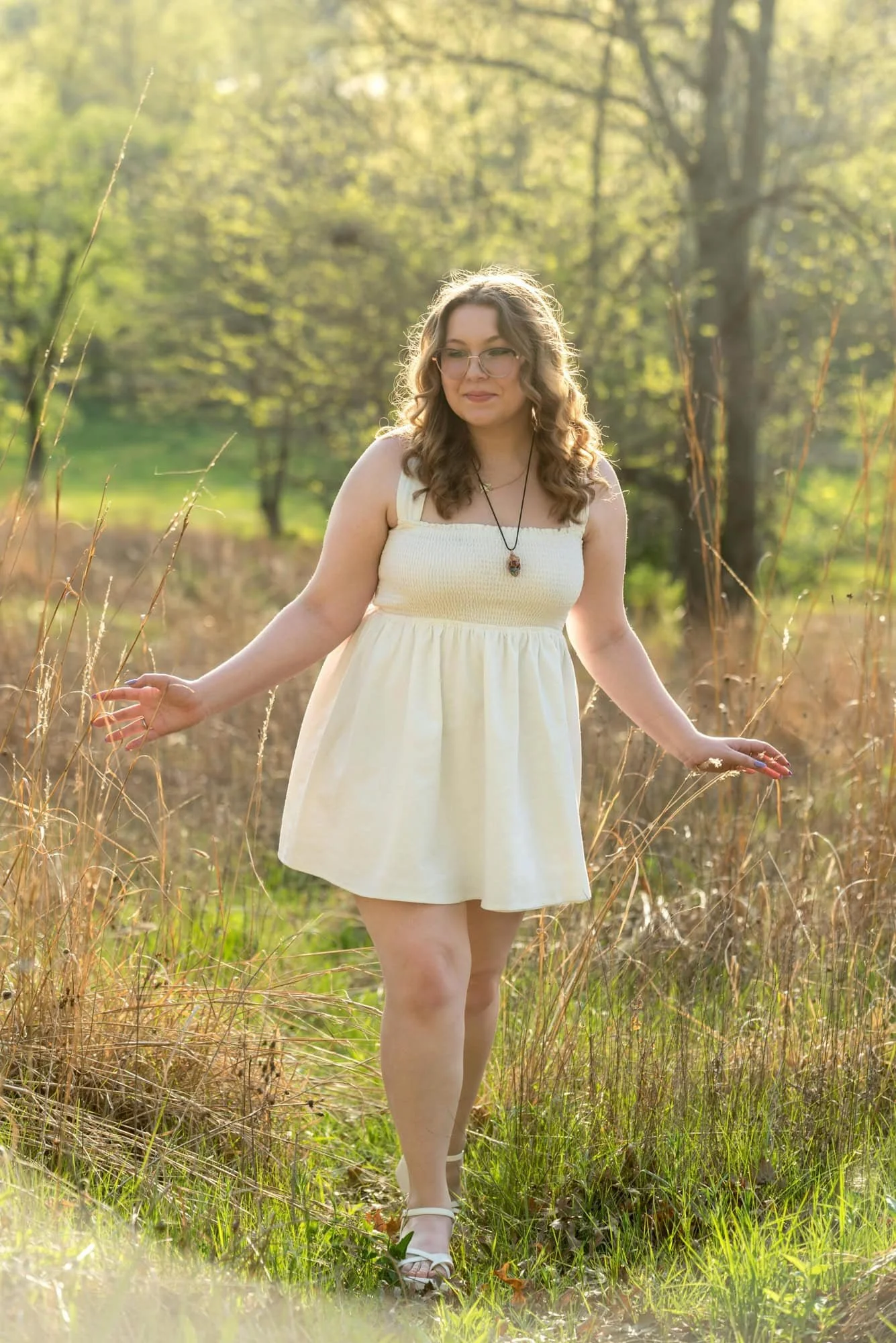 Walking through open grassy fields in Town and Country, a senior girl in a white dress smiles for a picture.