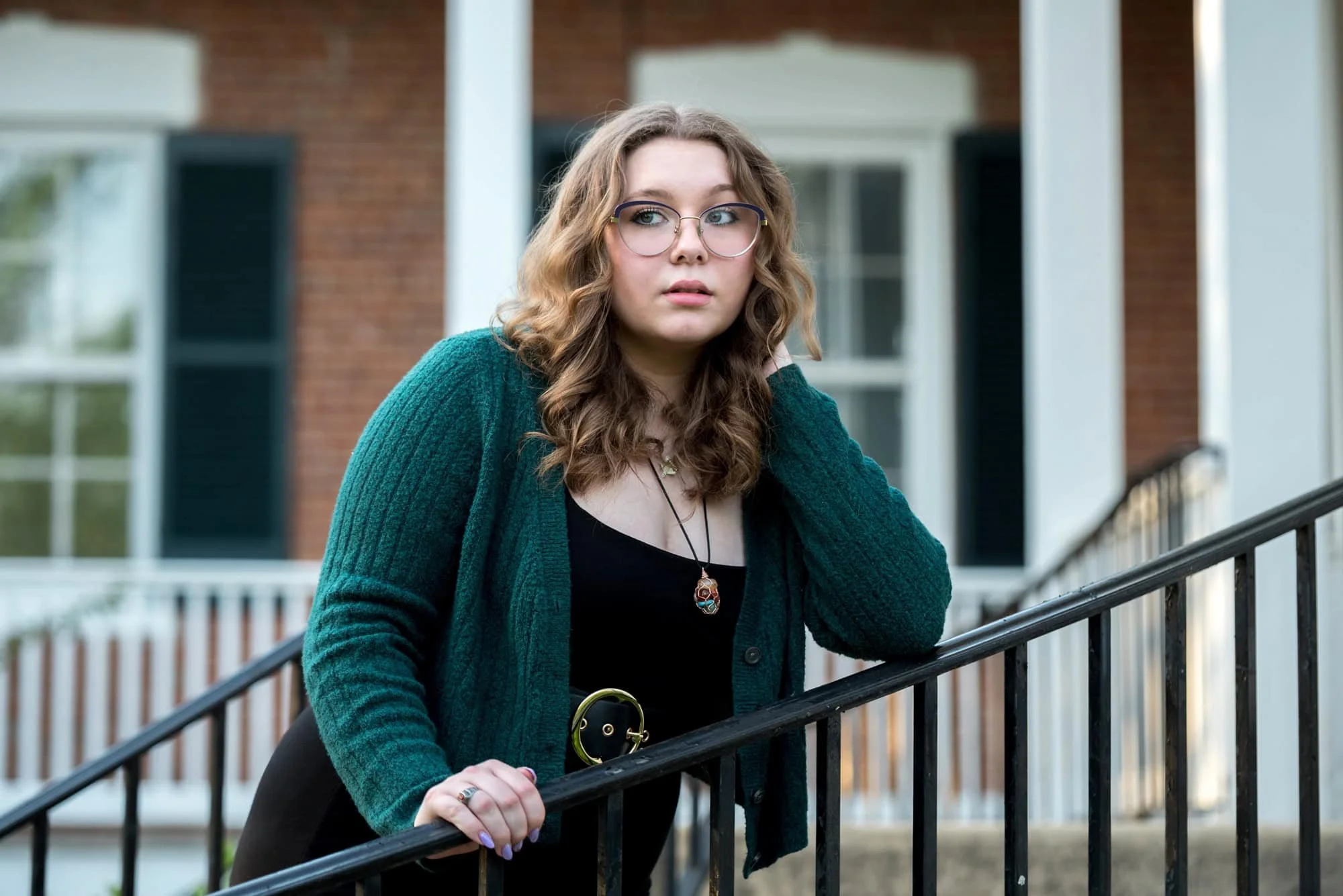 Posing on the steps of a historic home, a senior girl in a black dress and green cardigan looks off for a picture in West County.