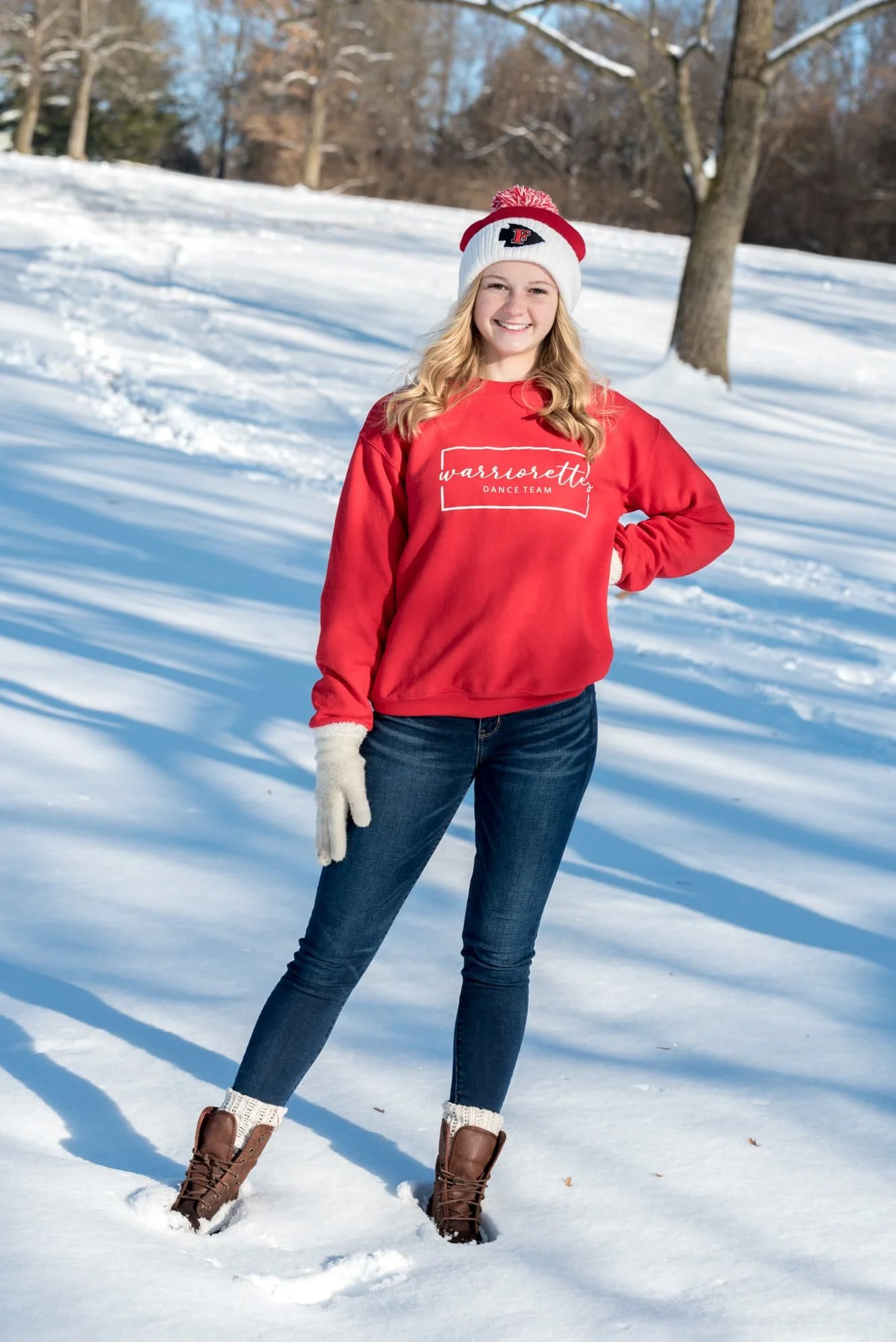 High school senior portrait by an Arnold MO senior photographer, showing a Fox High School student proudly posing in the snow at Suson Park wearing a red Fox Warriorettes sweatshirt, red beanie, blue jeans, white gloves, and brown boots.