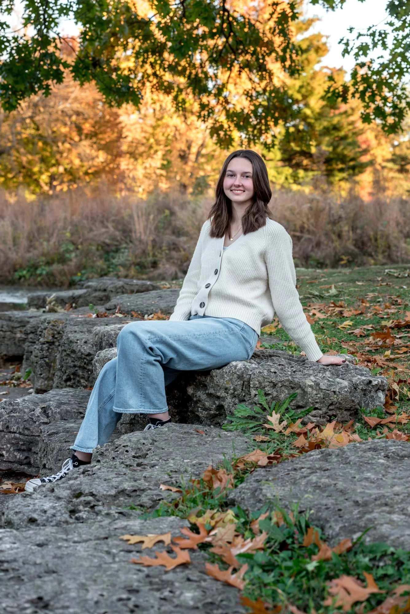 A high school senior girl wearing a white sweater and blue jeans leans back on the creek bed rocks taken by St. Louis senior photographer.