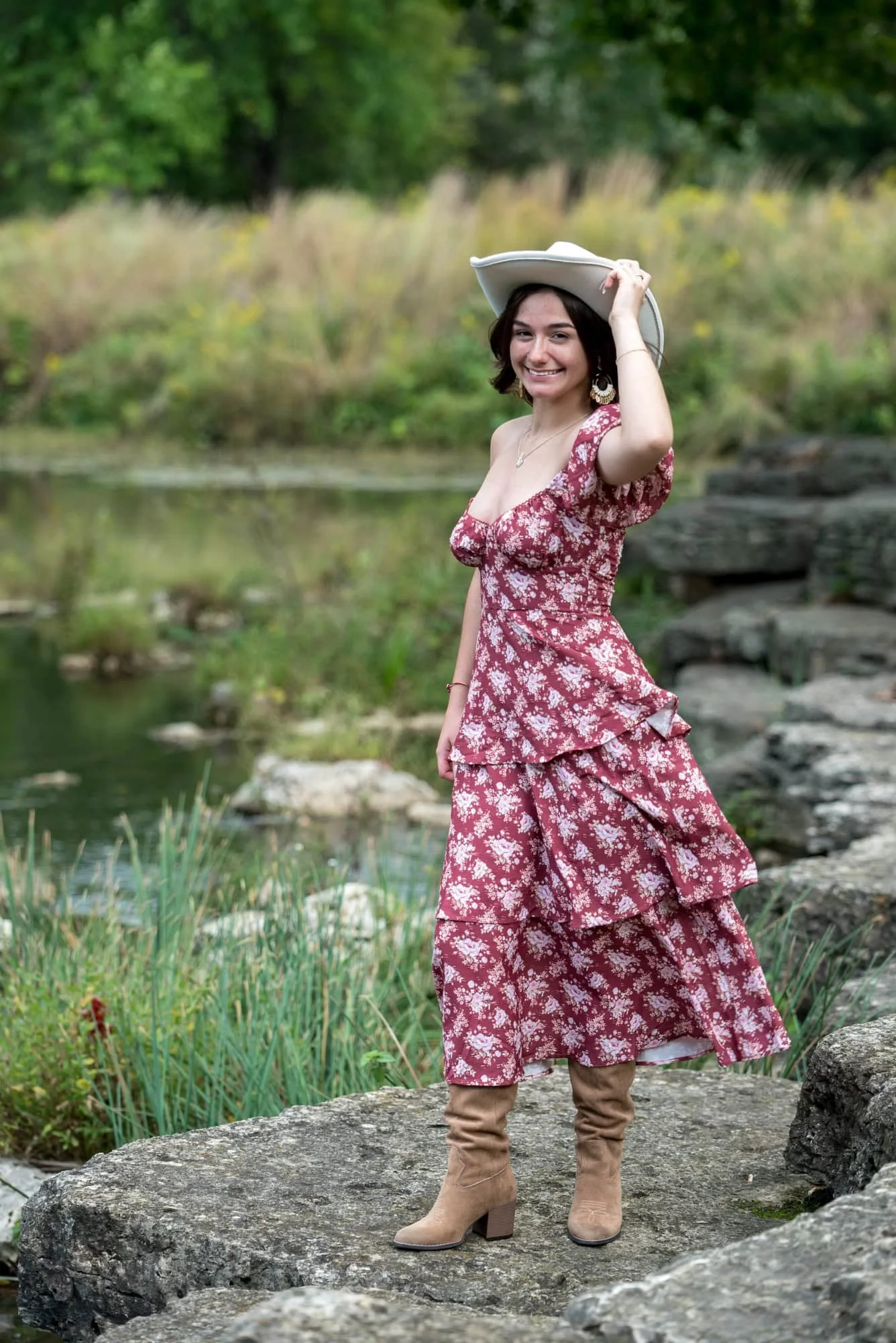 A picture captures a high school senior girl wearing a red dress, a white cowboy hat, and brown boots as she stands on a rock near a creek in Forest Park.